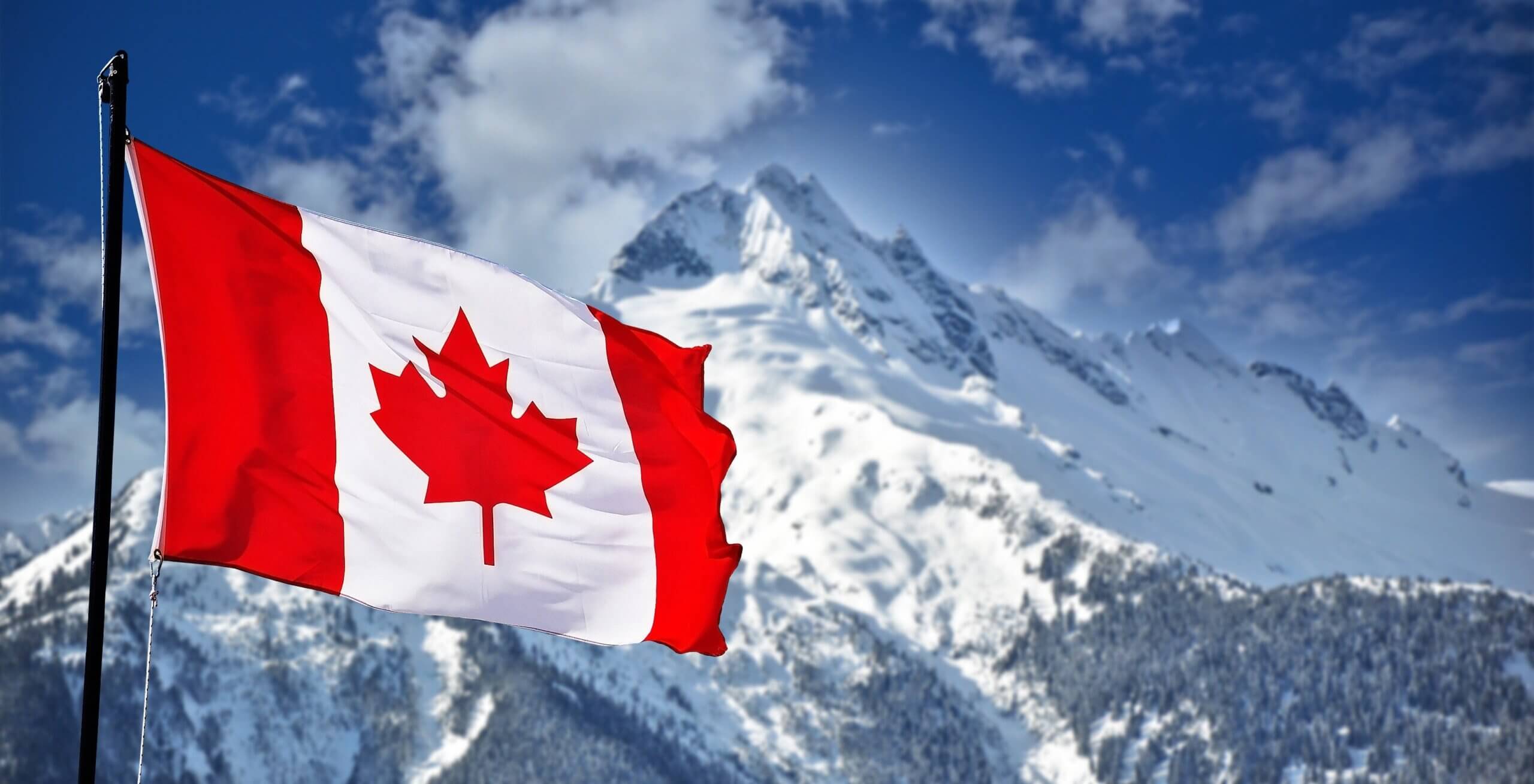 The Canadian flag, featuring a red maple leaf on a white background with red vertical stripes, waves in the foreground against a backdrop of snow-covered mountains under a clear blue sky.