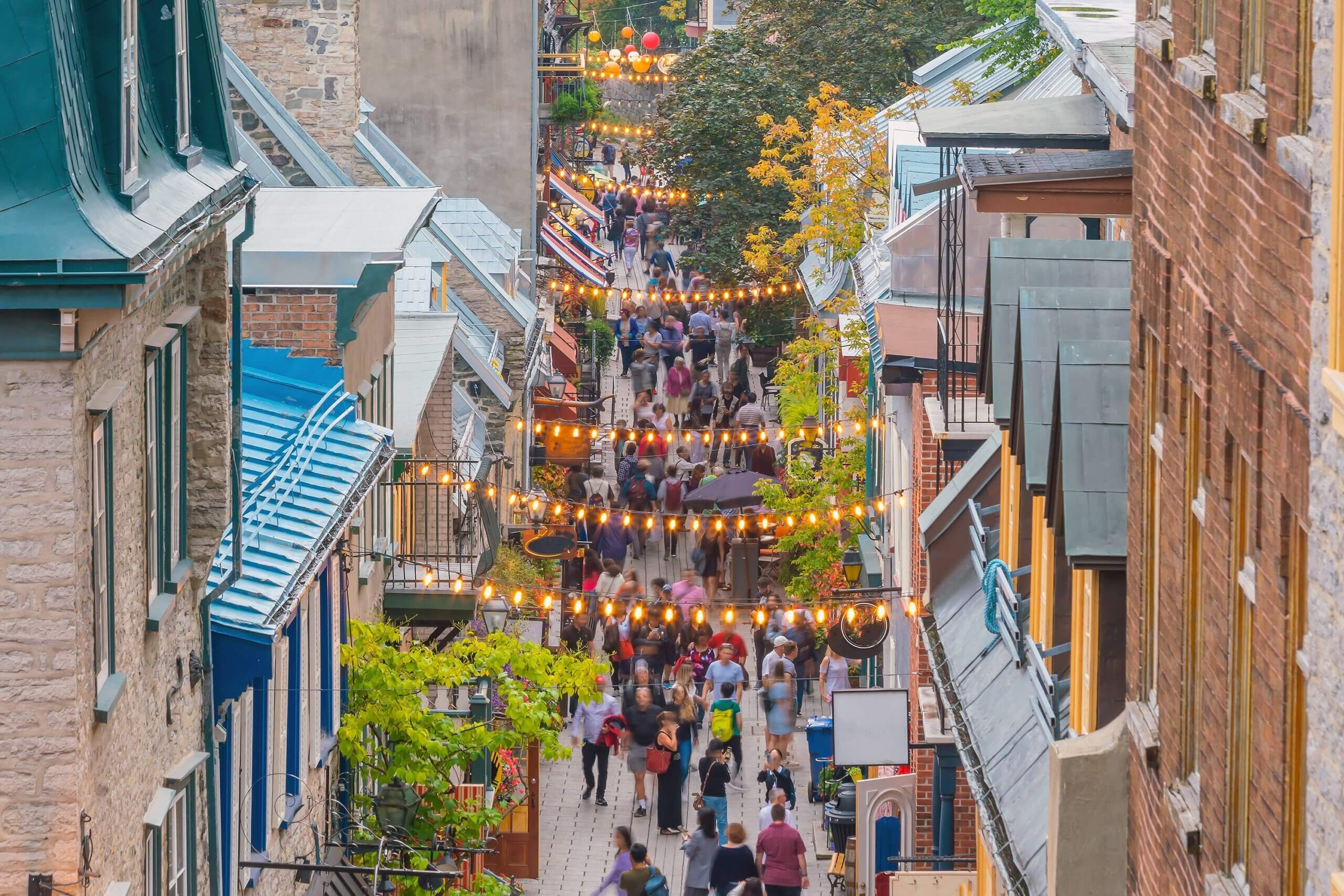 A lively, narrow street bustling with people. Strings of festive lights are hung between the buildings, which feature colorful roofs and masonry. Trees with autumn foliage add a touch of color to the scene.
