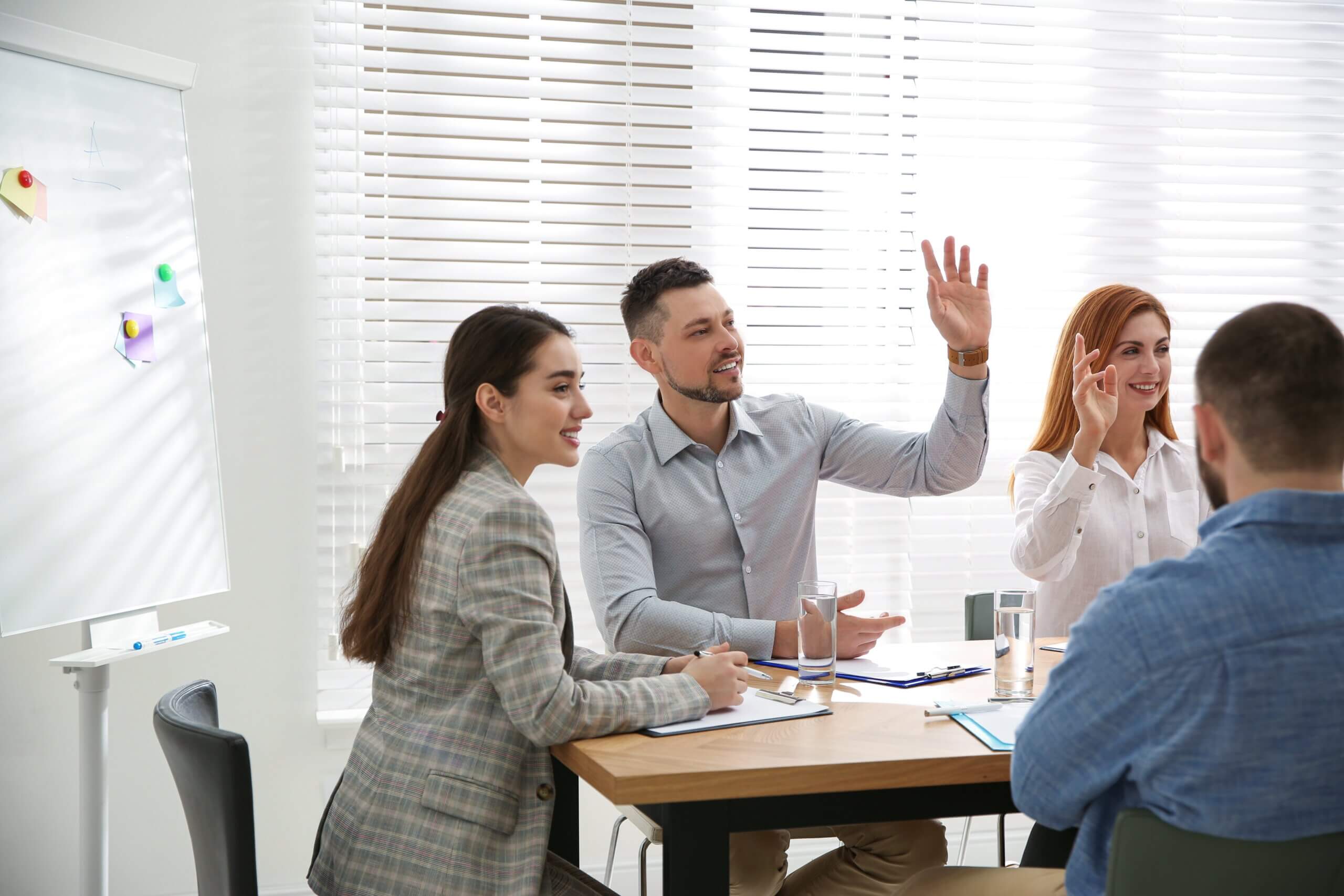 Four people sitting at a table in an office setting, engaged in a meeting. Two individuals have their hands raised. A whiteboard with colorful magnets is in the background. The room is well-lit with natural light from the blinds.