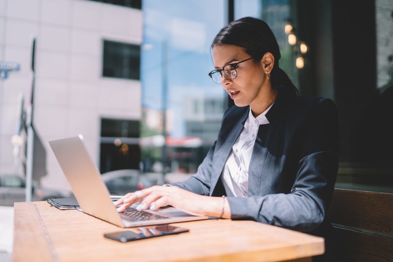 A woman wearing glasses and a suit is typing on a laptop at a wooden table in a modern office. A smartphone is placed beside her. She appears focused, and large windows reveal an urban backdrop.