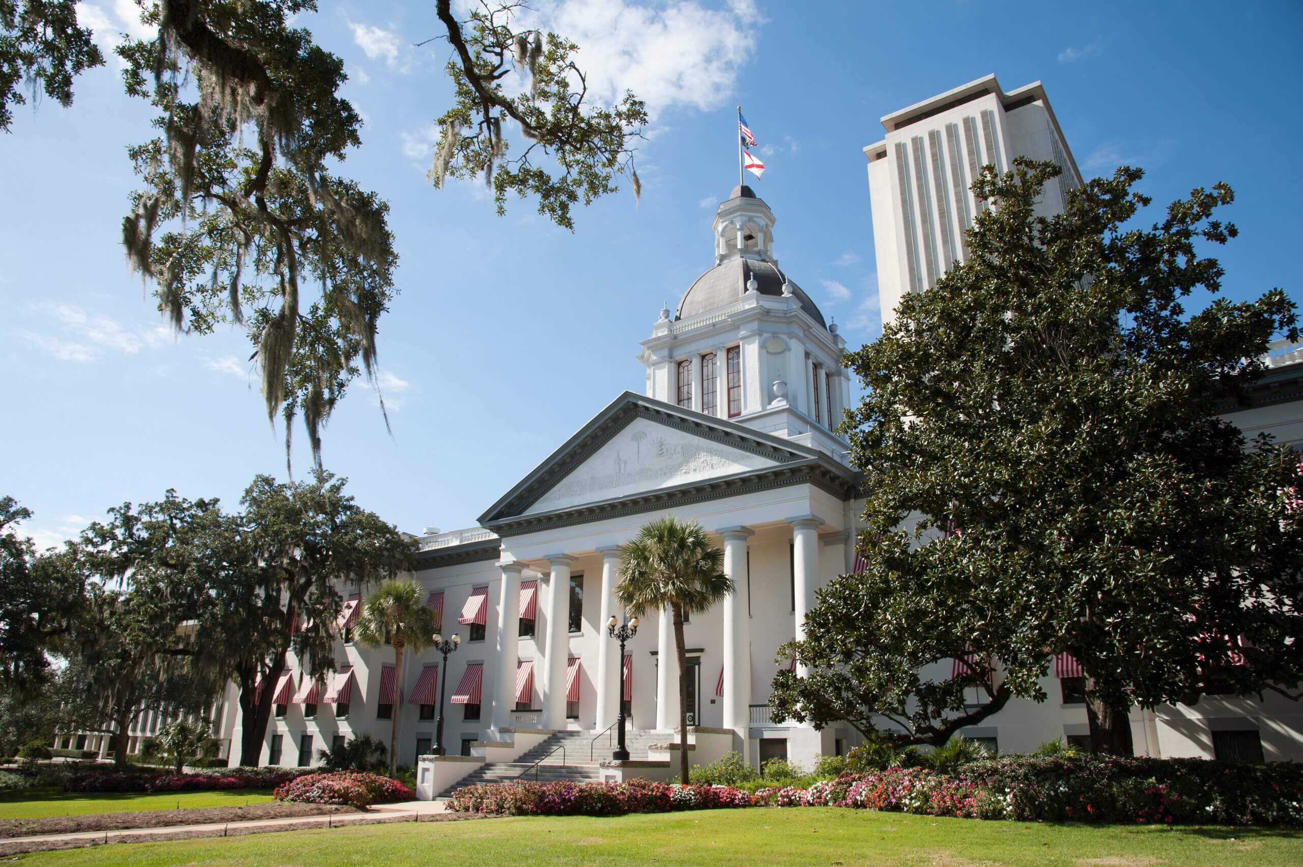 Historic building with white columns and a dome, surrounded by large trees and colorful flowers. Flags atop the dome and entrance, with a modern tower in the background under a blue sky.