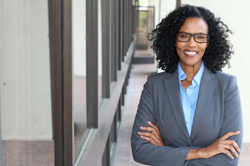 A woman with curly black hair and glasses smiles while standing with her arms crossed. She is wearing a gray blazer and a blue shirt. The background shows a corridor with large windows.