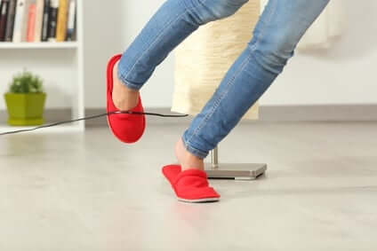 Person wearing red slippers and blue jeans trips over a black cord on a light-colored floor. A lamp and a green potted plant are visible in the background.