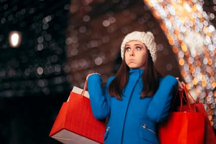 A woman in a blue coat and white knit hat holds multiple red shopping bags, looking up with a pout. She stands in front of a background filled with blurred, twinkling lights.