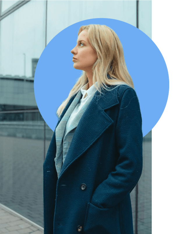 A person with long blonde hair, wearing a dark blue coat and light-colored shirt, stands in front of a reflective glass building. The background has a circular blue overlay.