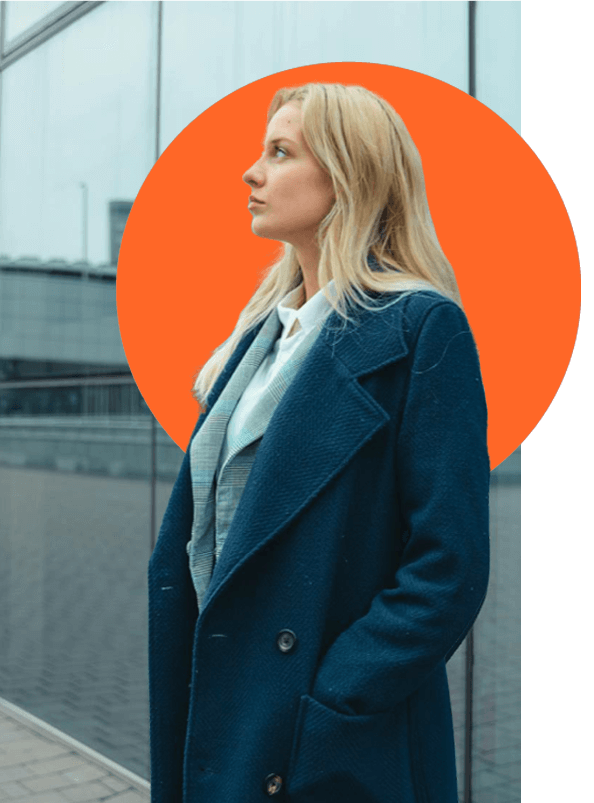 A woman with long blonde hair, wearing a blue coat over a light-colored outfit, stands in profile against a modern glass building. There's an orange circle backdrop behind her head.