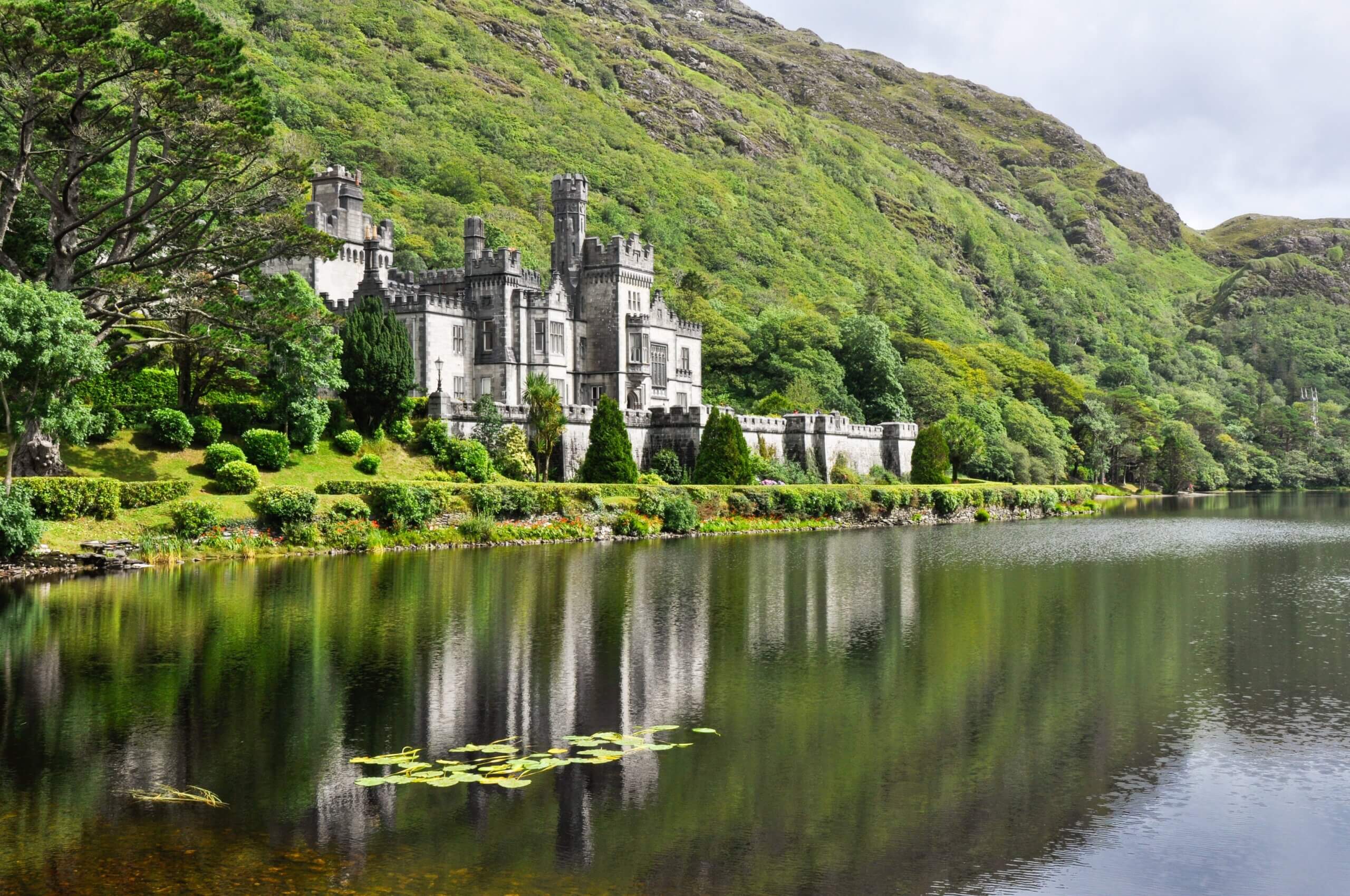 A historic castle with gray stone walls and multiple towers sits beside a tranquil lake. The building is surrounded by lush greenery, including trees and bushes. The landscape and building are reflected in the calm water.