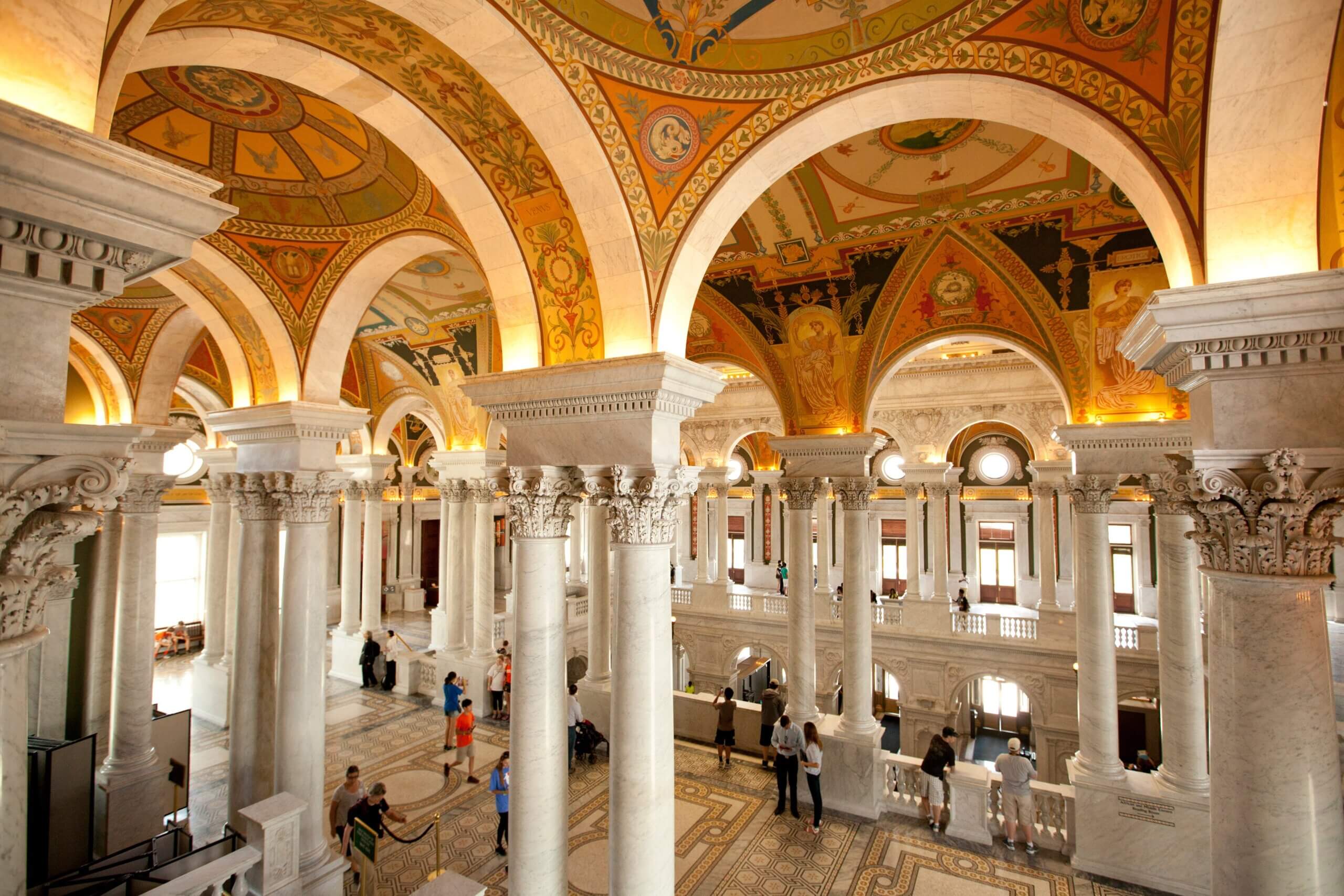 A grand, ornately decorated interior with marble columns and arches, intricate frescoes on the ceiling, and a spacious, patterned floor. People are walking and observing the architecture in the brightly lit hall.