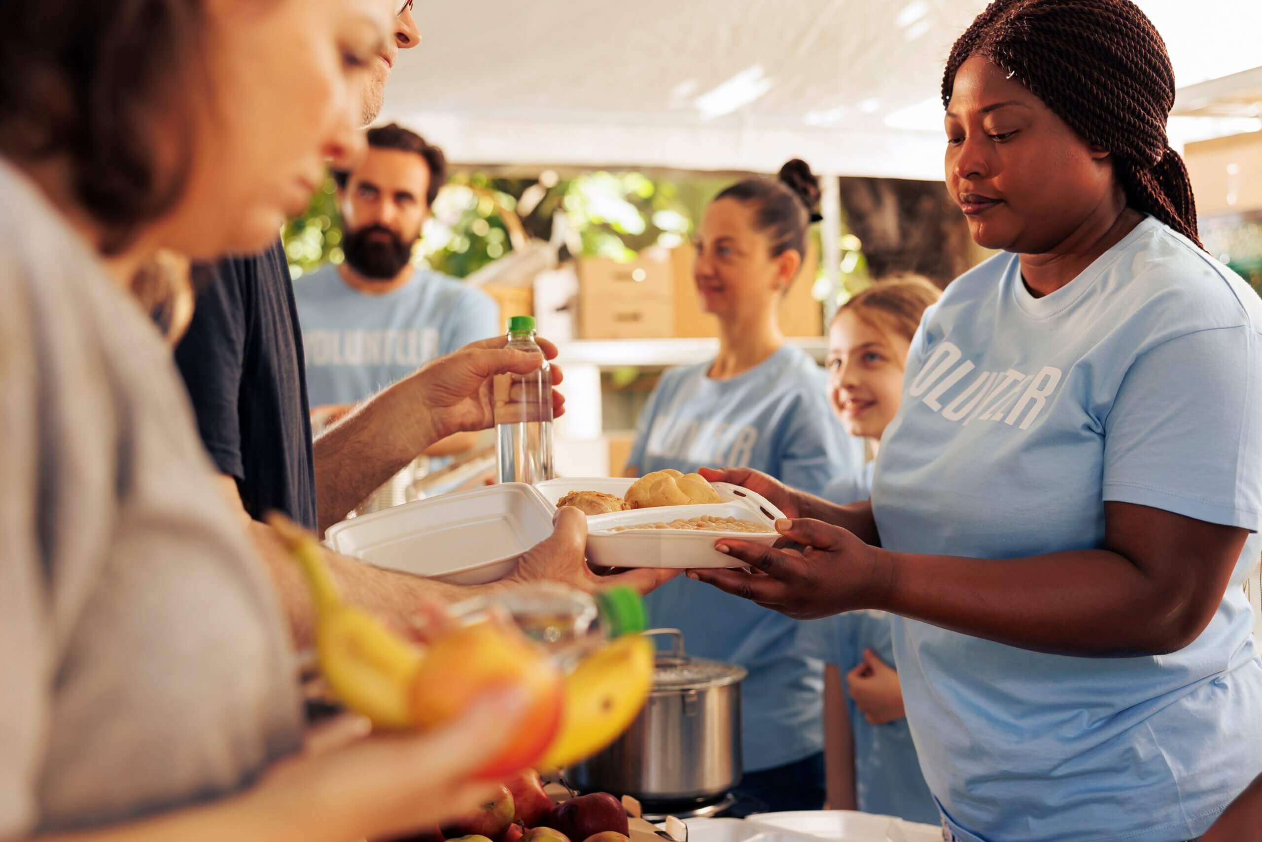 Volunteers in blue shirts distribute food at an outdoor event. A woman hands out a meal in a takeout container. Fruits and other food items are visible on the table. People of various ages participate, with trees in the background.