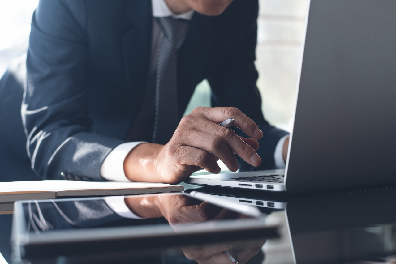 A person in a suit is leaning over a laptop, using a pen to navigate the touchpad. A notebook and tablet are on the table, with the background softly blurred, conveying a focused work environment.