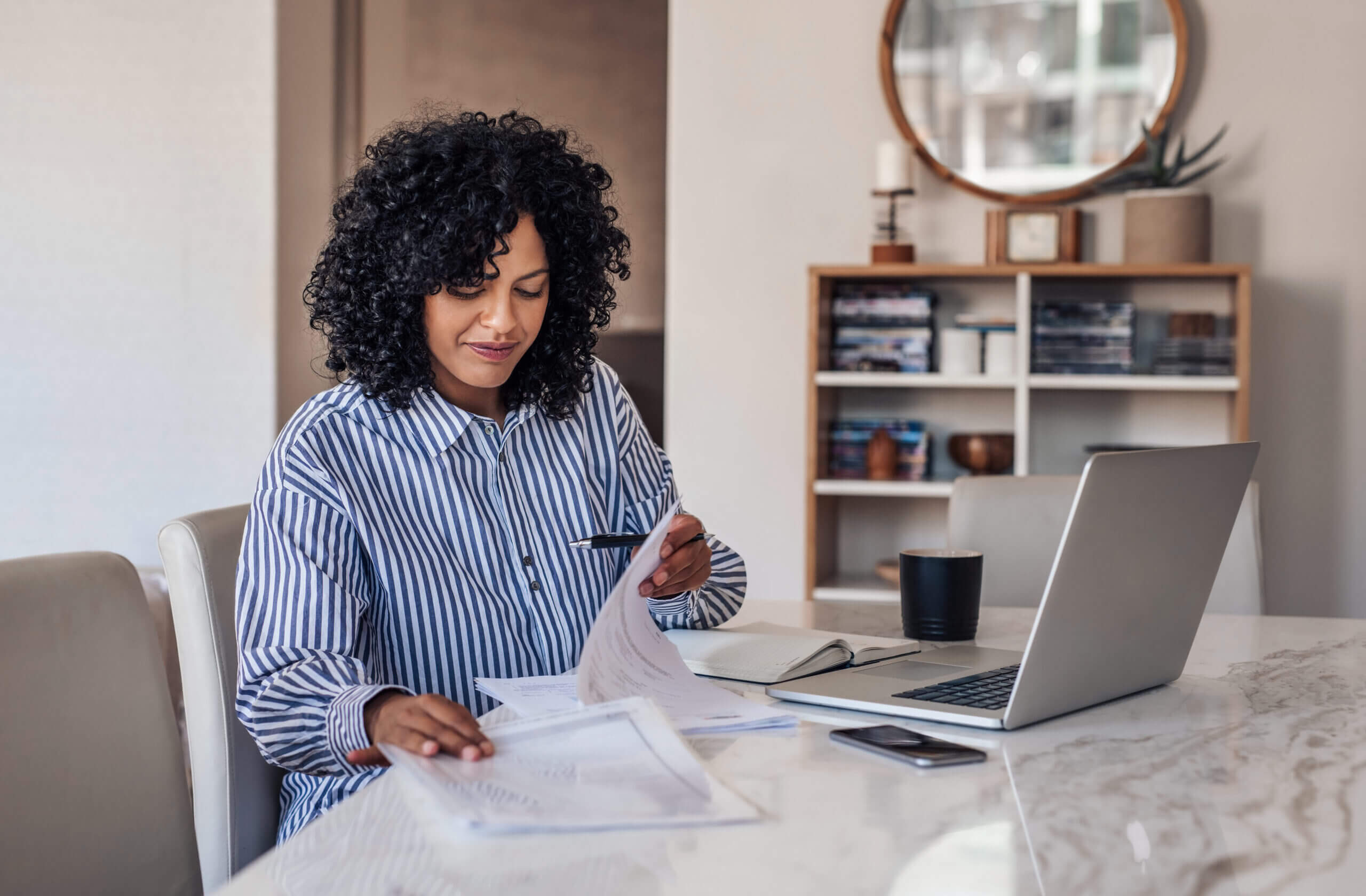 Woman with curly hair reviews papers at a table with a laptop, phone, and mug. She's wearing a striped shirt in a well-lit room with a bookshelf and a round mirror in the background.