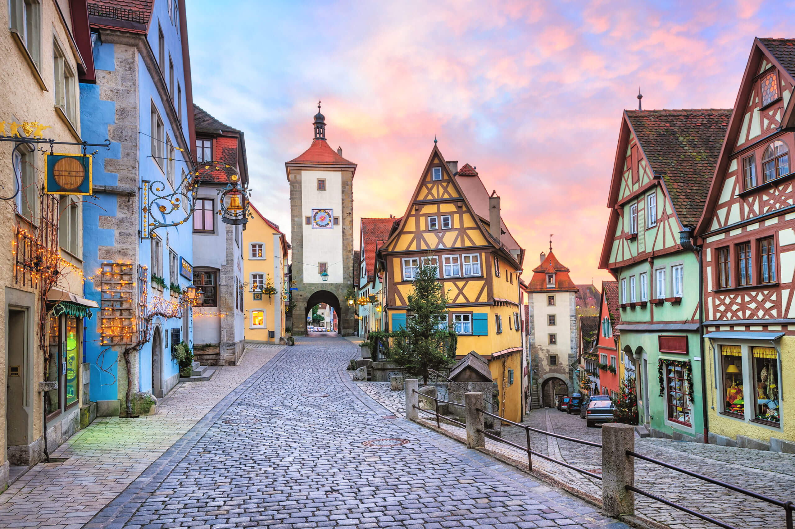 Cobblestone street in Rothenburg ob der Tauber, Germany, lined with colorful medieval buildings at sunset. A central clock tower is in the background, and storefronts with warm lights are on the left.