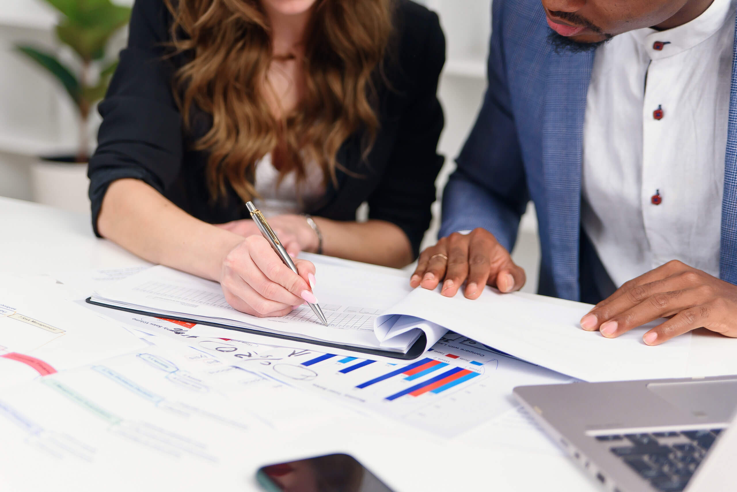 Two people working together at a desk. One is writing on a document, while the other is pointing at charts and graphs. A laptop and smartphone are nearby. The scene suggests collaboration or data analysis.