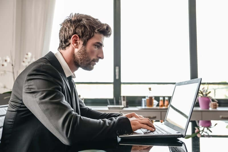 A man in a black suit is sitting at a desk, typing on a laptop. He has short hair and a beard. The background features large windows with partially drawn curtains and some indoor plants on the windowsill.