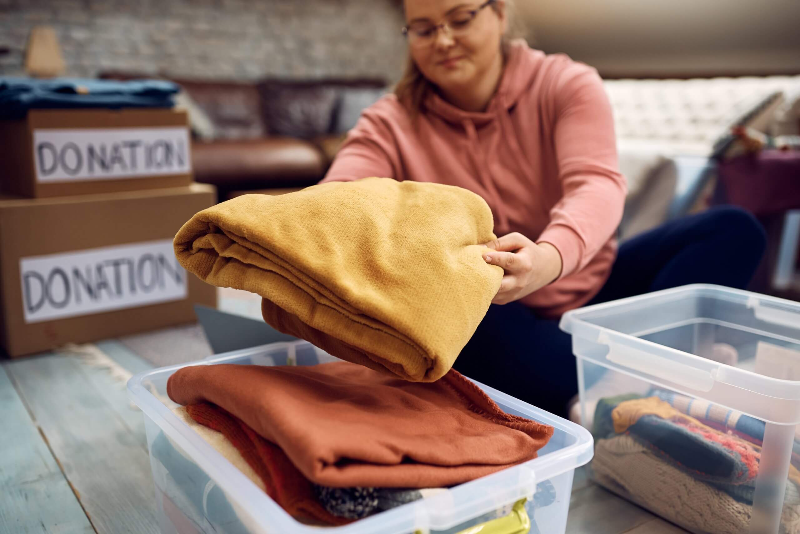 A person wearing glasses and a pink hoodie is sitting on the floor, folding a yellow blanket into a clear plastic storage bin. Nearby, there are boxes labeled "Donation" filled with clothes. The background shows a couch and more storage bins.