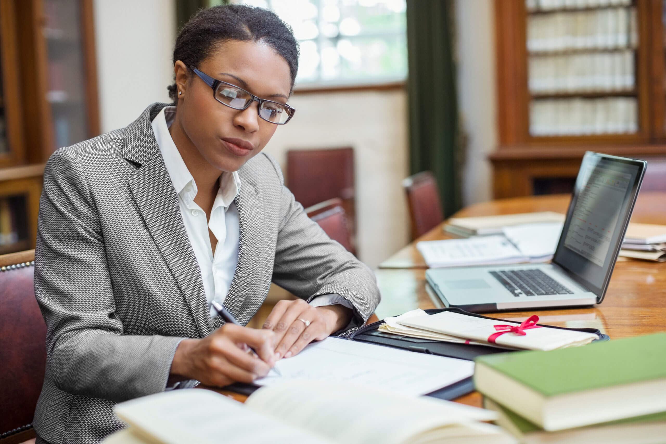 A woman in a suit and glasses is writing at a wooden table surrounded by books and a laptop in a library. She appears focused and engaged in her work. Shelves filled with books are visible in the background.