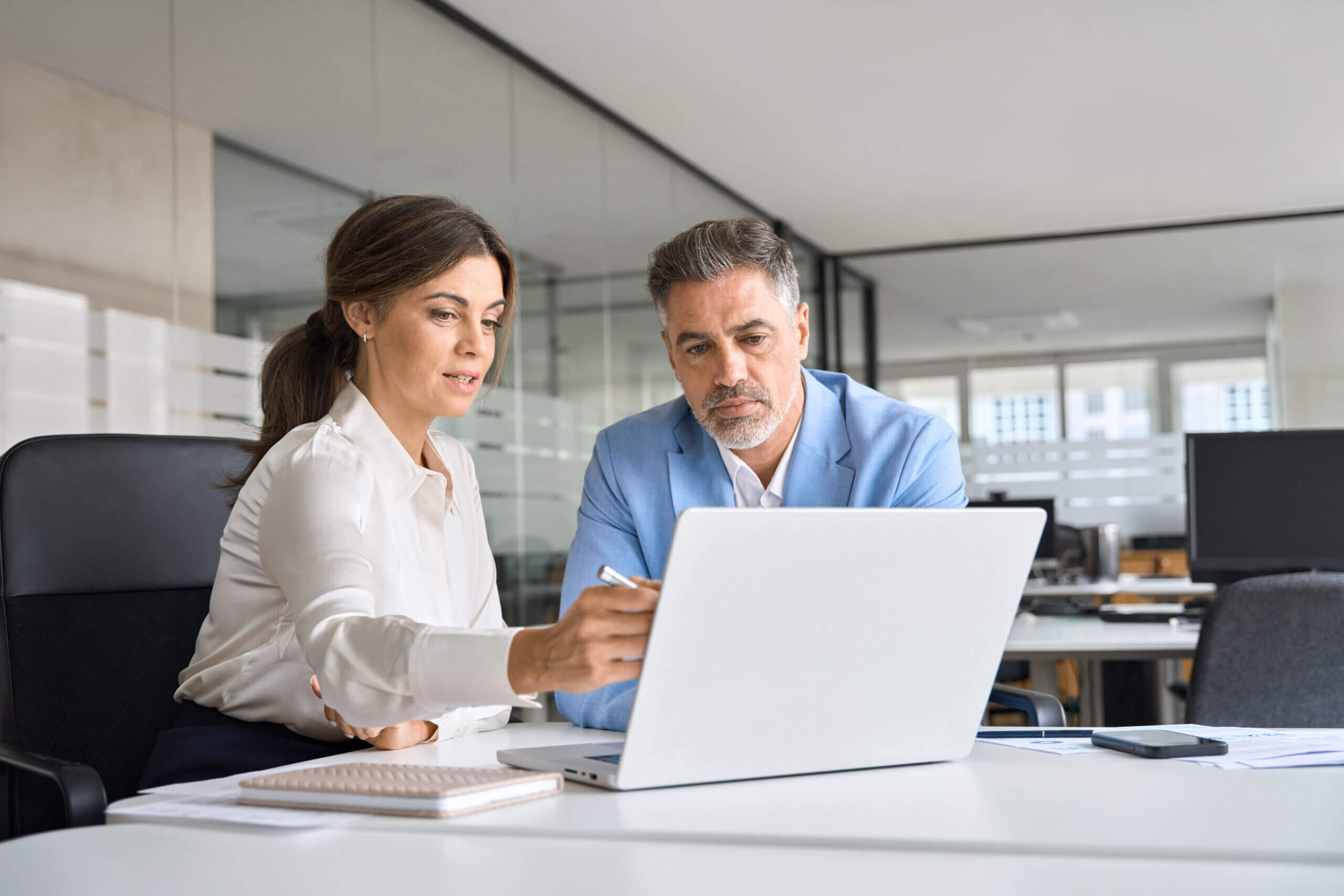 A woman and a man are seated at a desk in an office, looking at a laptop screen. The woman is pointing at the screen, explaining something to the man. Papers and a notebook are on the desk. The office has large windows and modern decor.