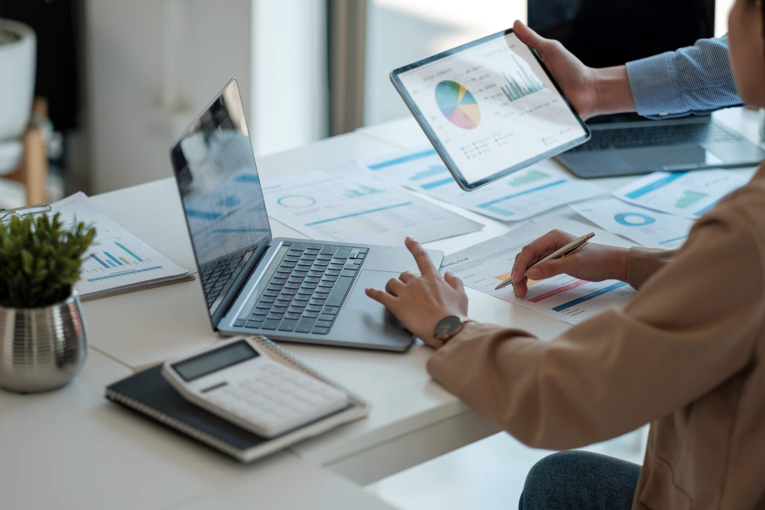 People working at a table with a laptop, tablet, papers, and a calculator. Charts and graphs are displayed on the devices and documents. One person points at a graph on paper, while another holds up a tablet showing a pie chart.