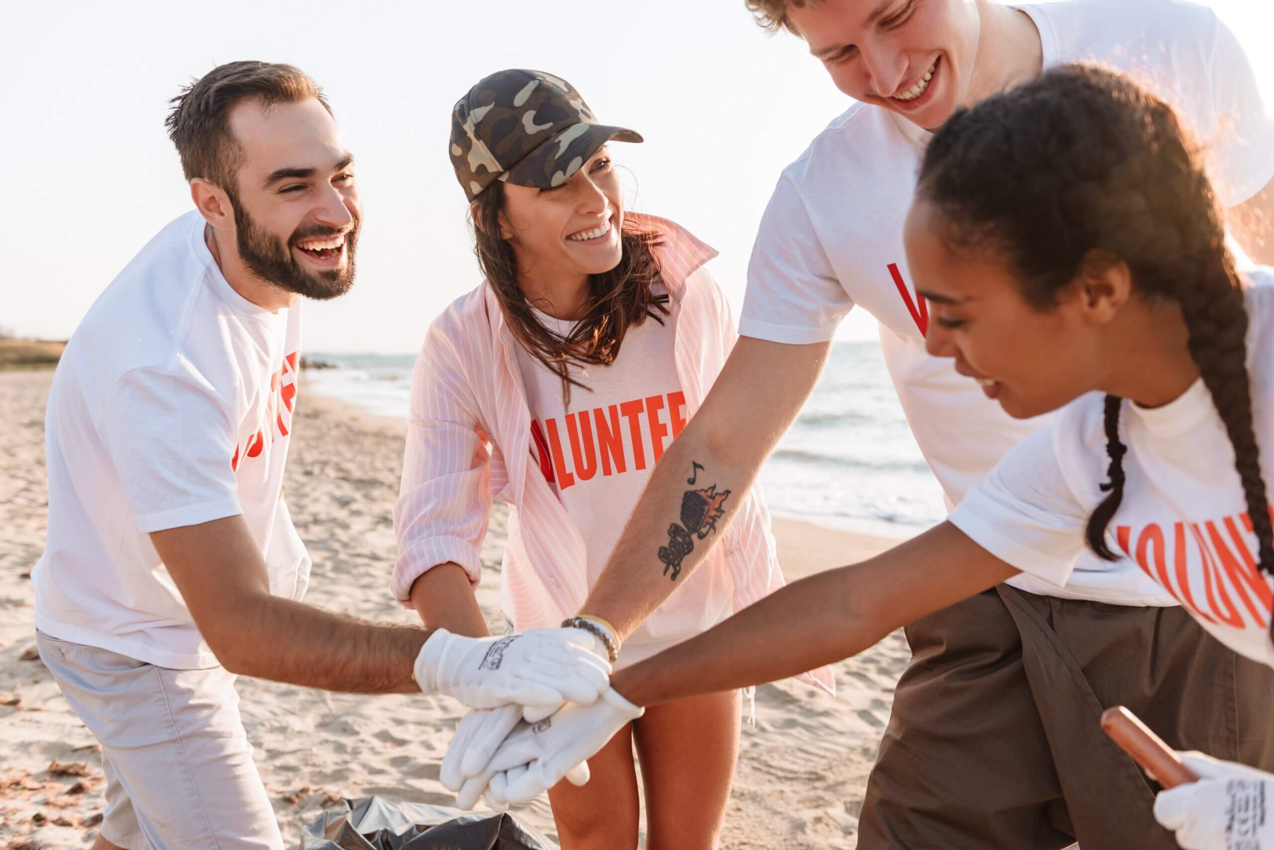 A group of four people wearing "Volunteer" t-shirts are standing on a beach, smiling and stacking their hands together in a gesture of teamwork. The beach and ocean are visible in the background.