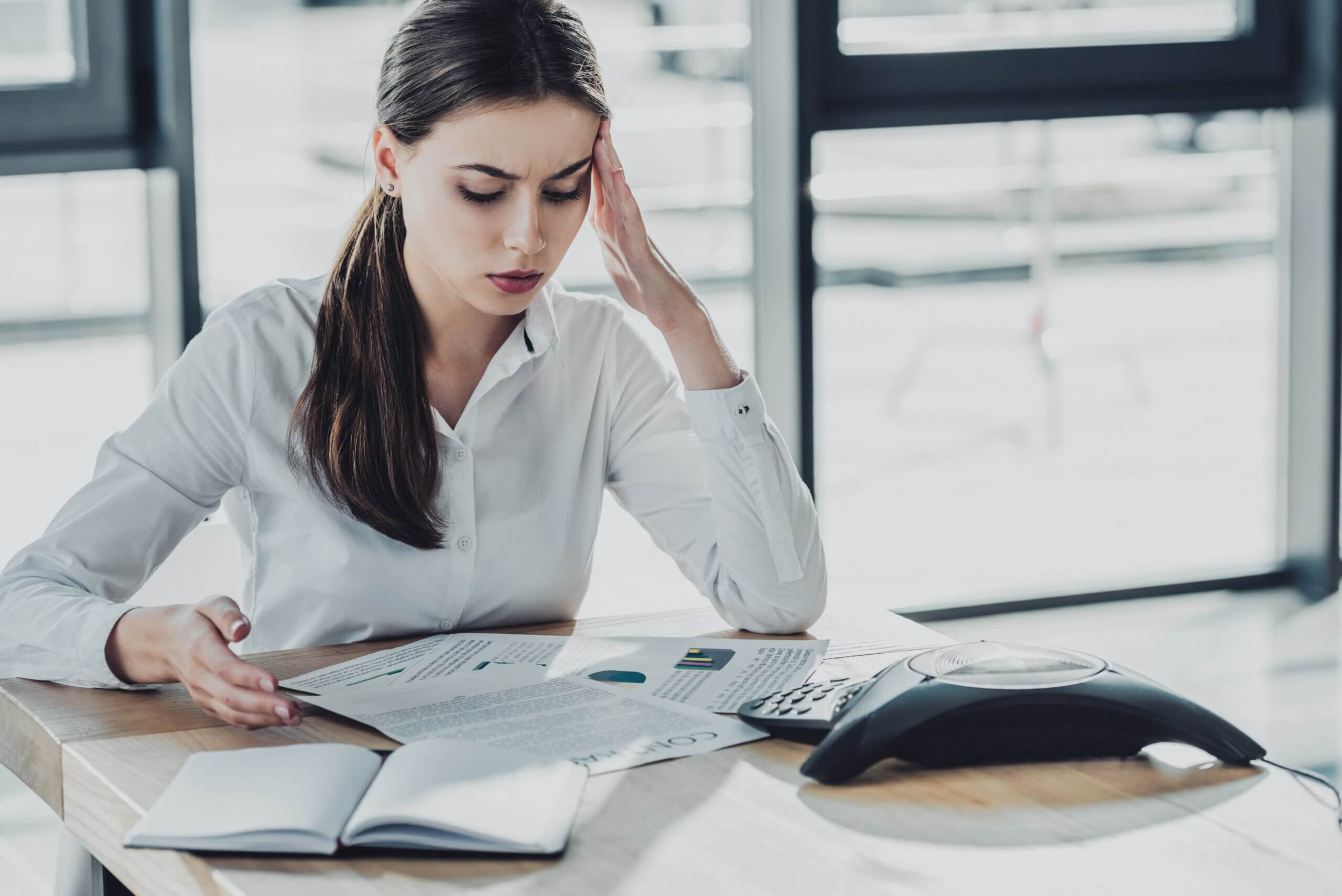 A woman in a white blouse sits at a desk with her head propped on one hand, looking at documents. An open notebook and a phone are on the table. She appears to be focused and concerned.