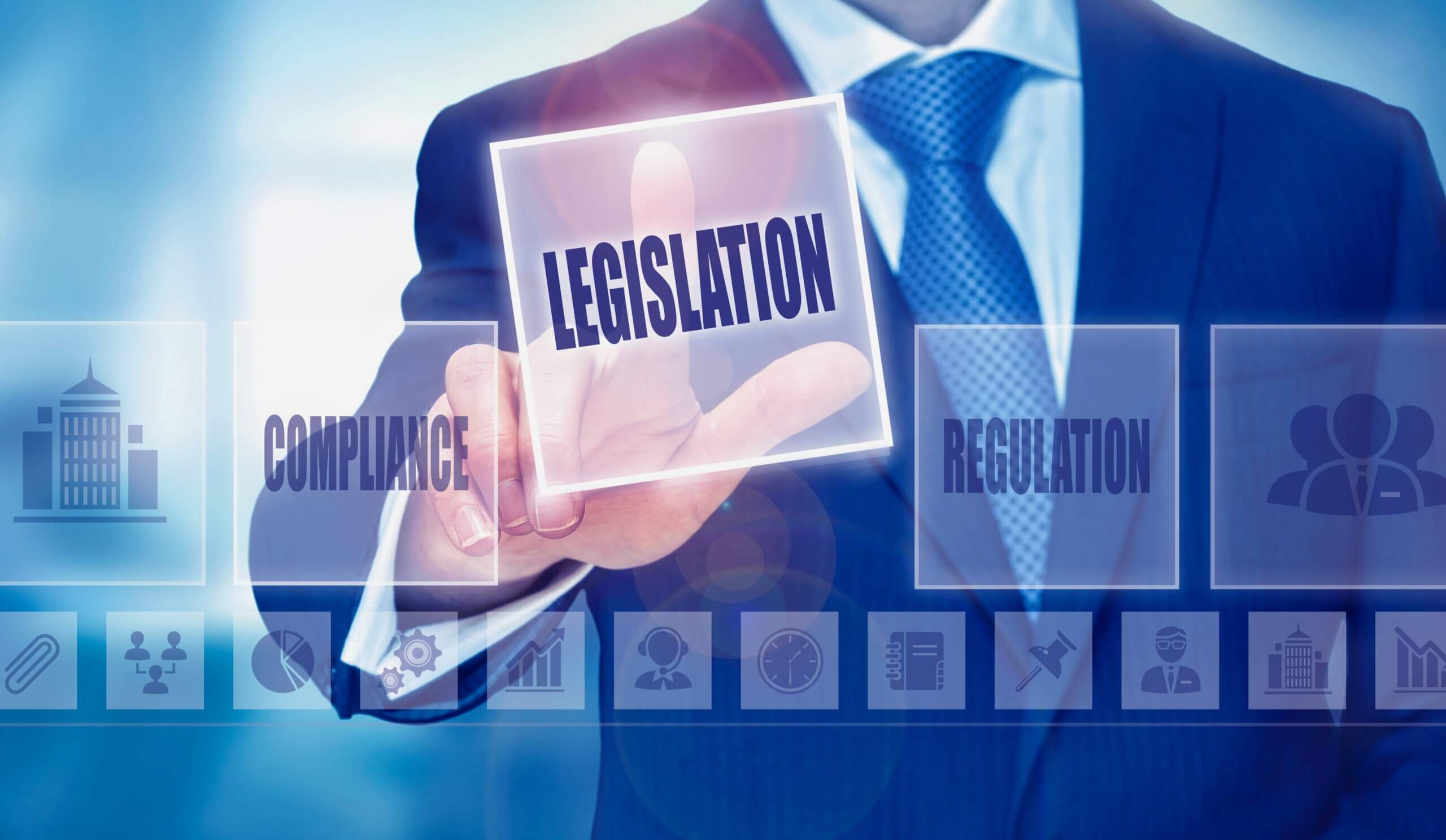 A person in a suit touches a transparent screen displaying the word "LEGISLATION" prominently. Other words visible on the screen include "COMPLIANCE" and "REGULATION," along with icons representing various business-related concepts.