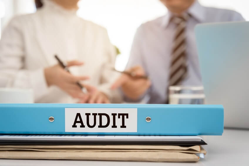 A blue binder labeled "Audit" is in the foreground. In the background, two people are in discussion, sitting at a desk with documents and a laptop. Their faces are not visible.