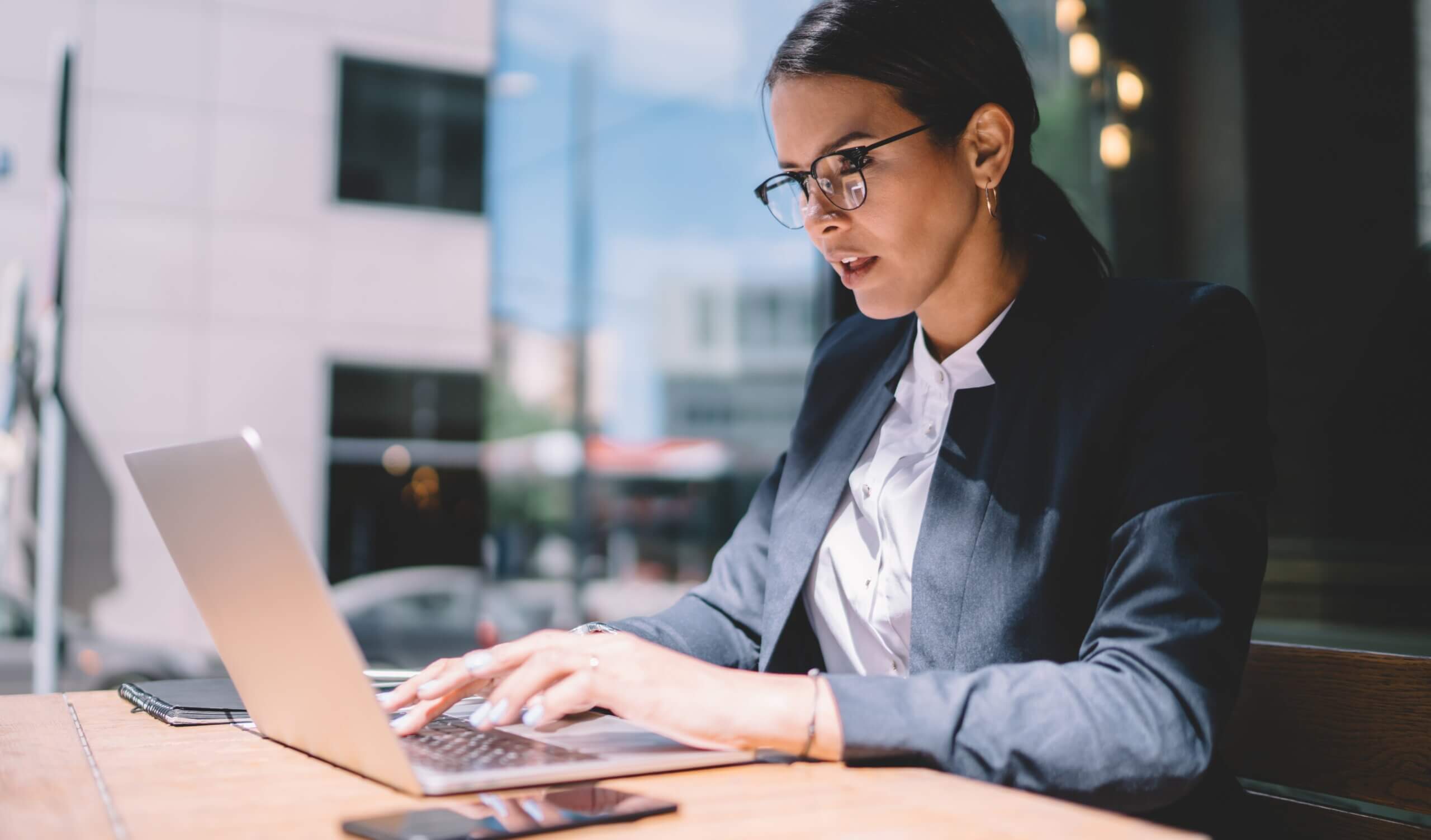 A woman in a business suit is working on a laptop at an outdoor table. She is focused, with a smartphone and a notepad next to her. The background shows a blurred view of urban buildings and light traffic.