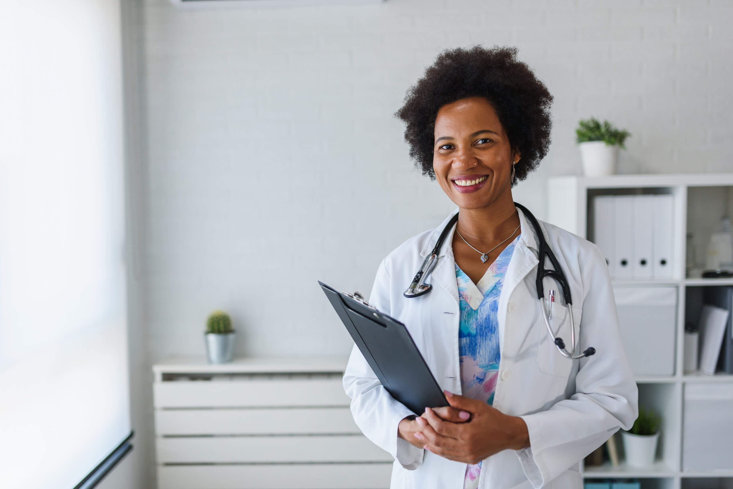 A smiling doctor with curly hair wearing a white coat and stethoscope holds a clipboard. She stands in a well-lit office with a white brick wall, shelves with folders, and small plants in the background.