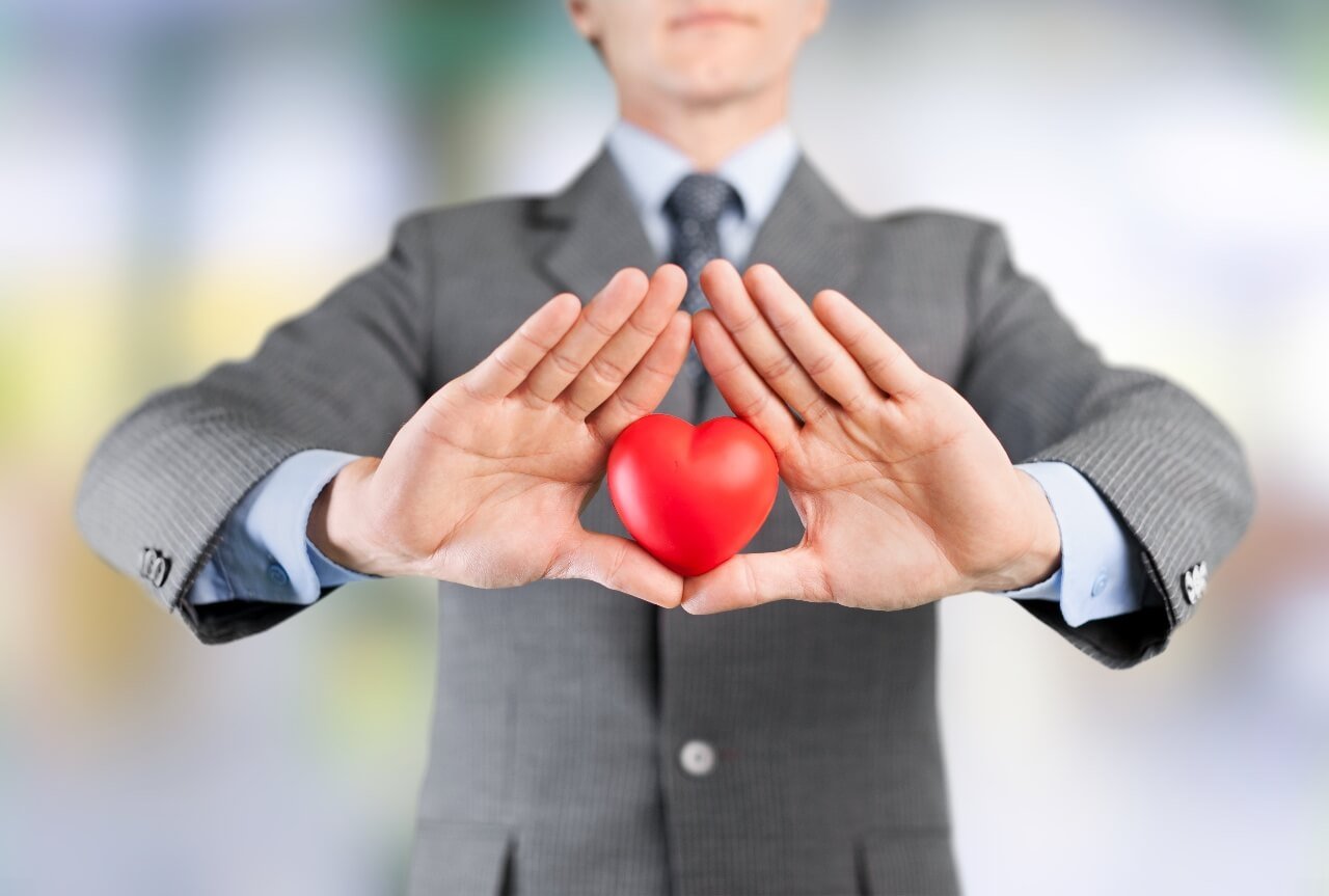 A person in a suit holds a small red heart between both hands, framing it with fingertips touching. The background is blurred and colorful.