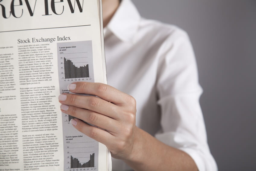 A person in a white shirt holds a newspaper open, showing a section titled "Stock Exchange Index" with text and two small line graphs. The person's face is not visible. The background is a plain gray.