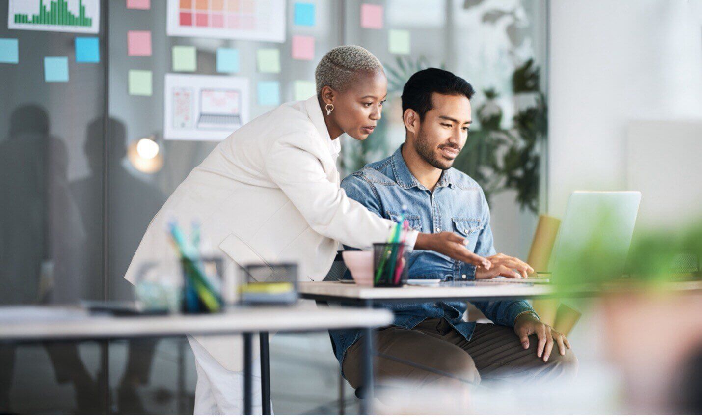 A woman and a man work together in an office. The woman, wearing a white blazer, leans over and gestures at the man's laptop screen. The man, in a blue shirt, sits focused. Sticky notes and charts are visible in the background.