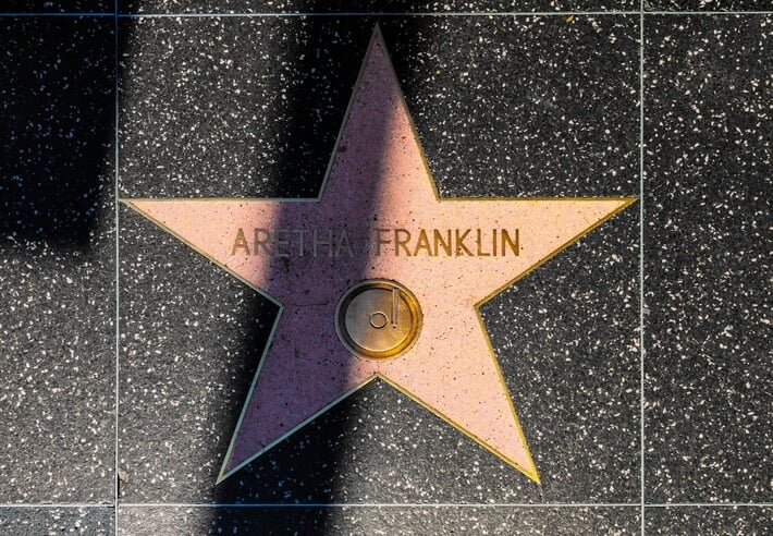 A pink star with a brass border and the name "ARETHA FRANKLIN" is embedded in a speckled black sidewalk. A circular symbol with a camera motif is beneath the name. A shadow partially covers the star.