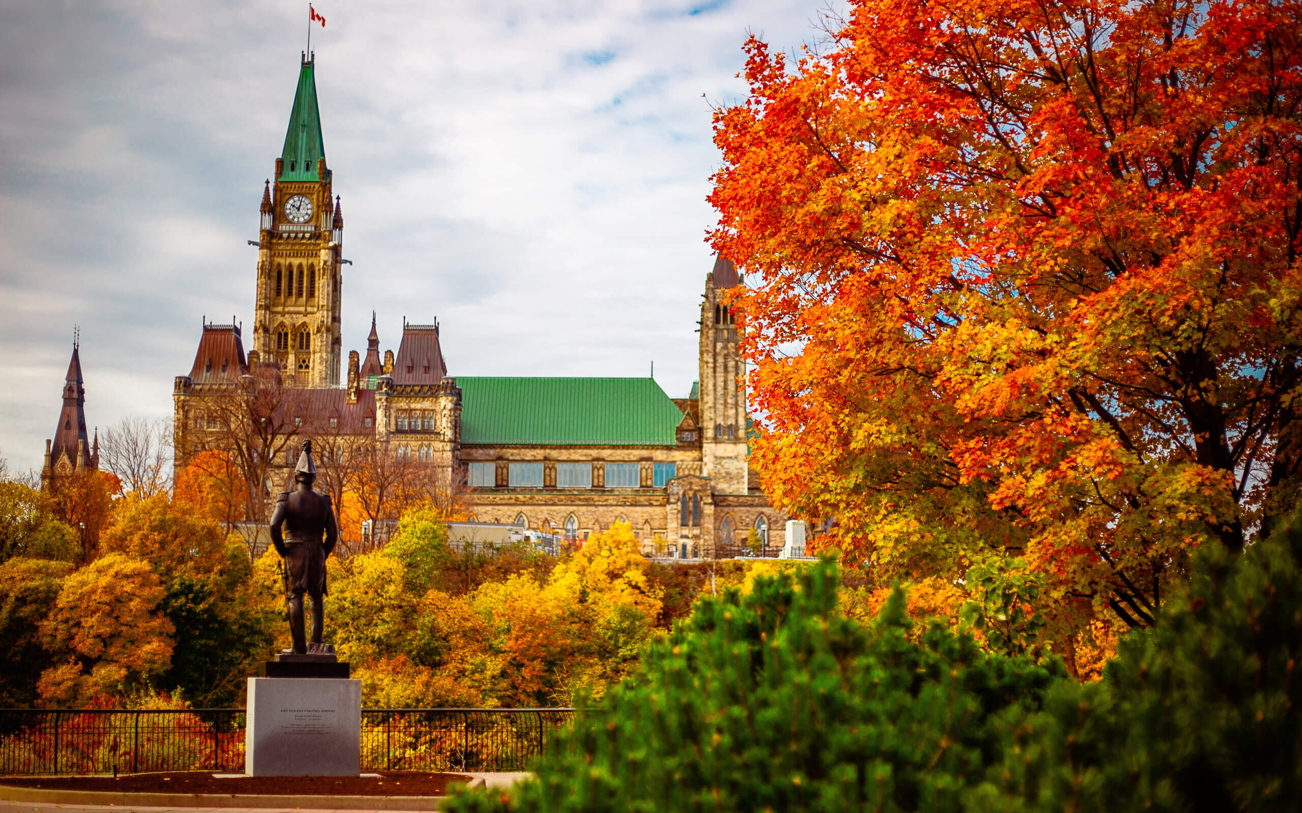Parliament Hill in Ottawa, Canada, is seen with vibrant autumn foliage. The Peace Tower is prominent against a cloudy sky. A statue stands in the foreground surrounded by colorful trees, adding a vibrant contrast.