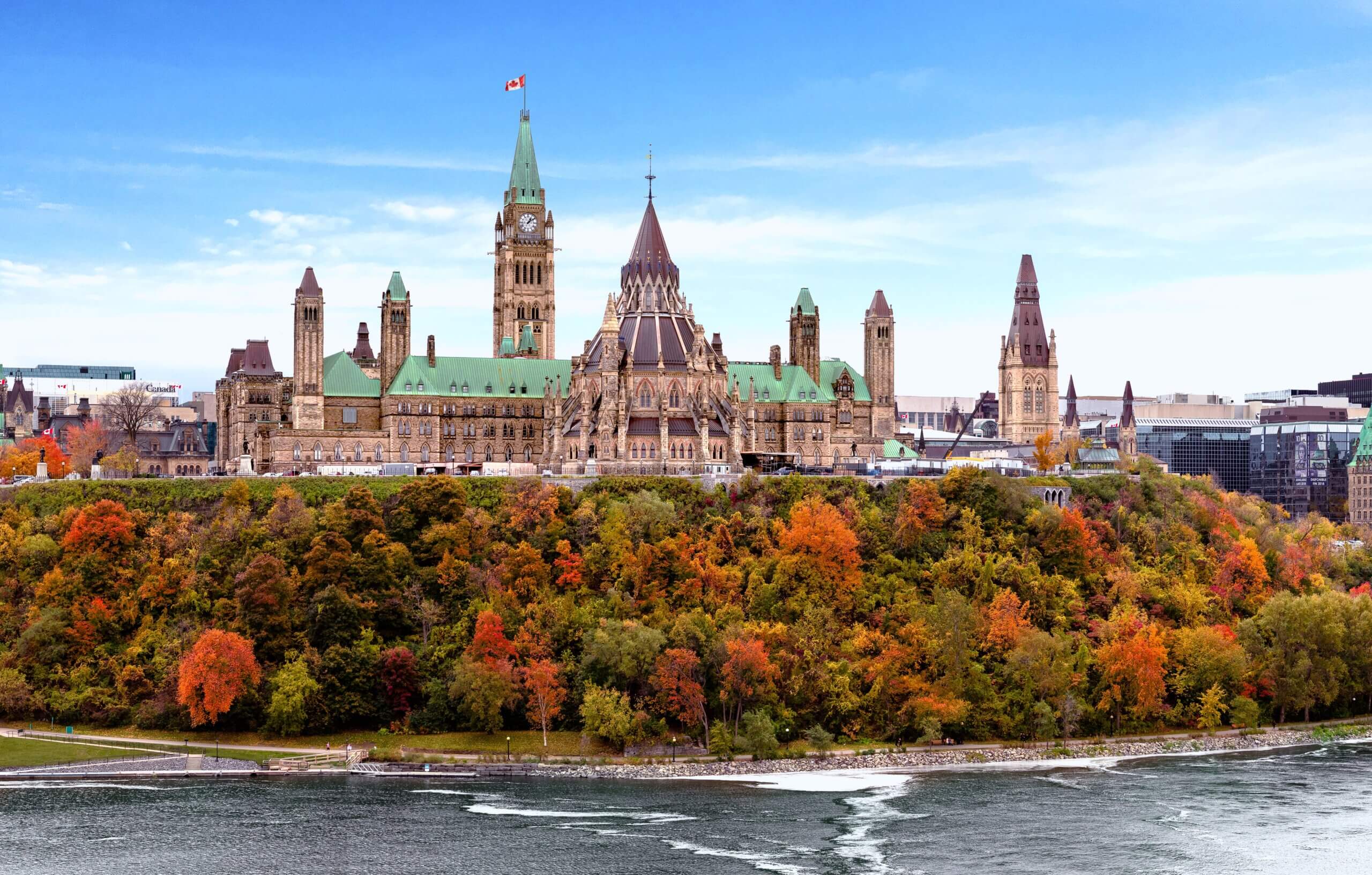 View of the Canadian Parliament buildings in Ottawa during autumn. The gothic-style architecture is surrounded by vibrant autumn foliage. The Ottawa River is in the foreground under a clear blue sky.