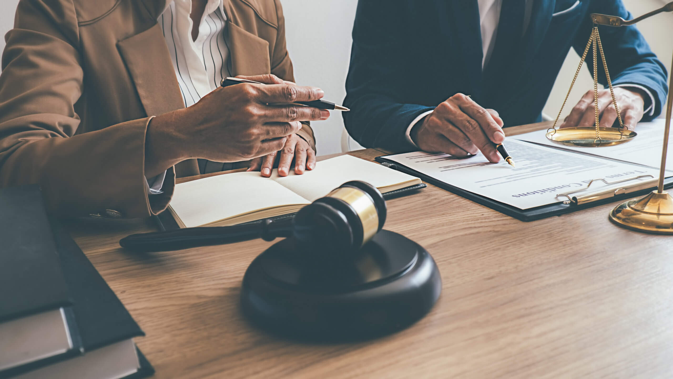 Two individuals in formal attire are seated at a desk, reviewing legal documents. A wooden gavel and scales of justice are seen on the desk. One person points with a pen, while the other takes notes in a notebook.
