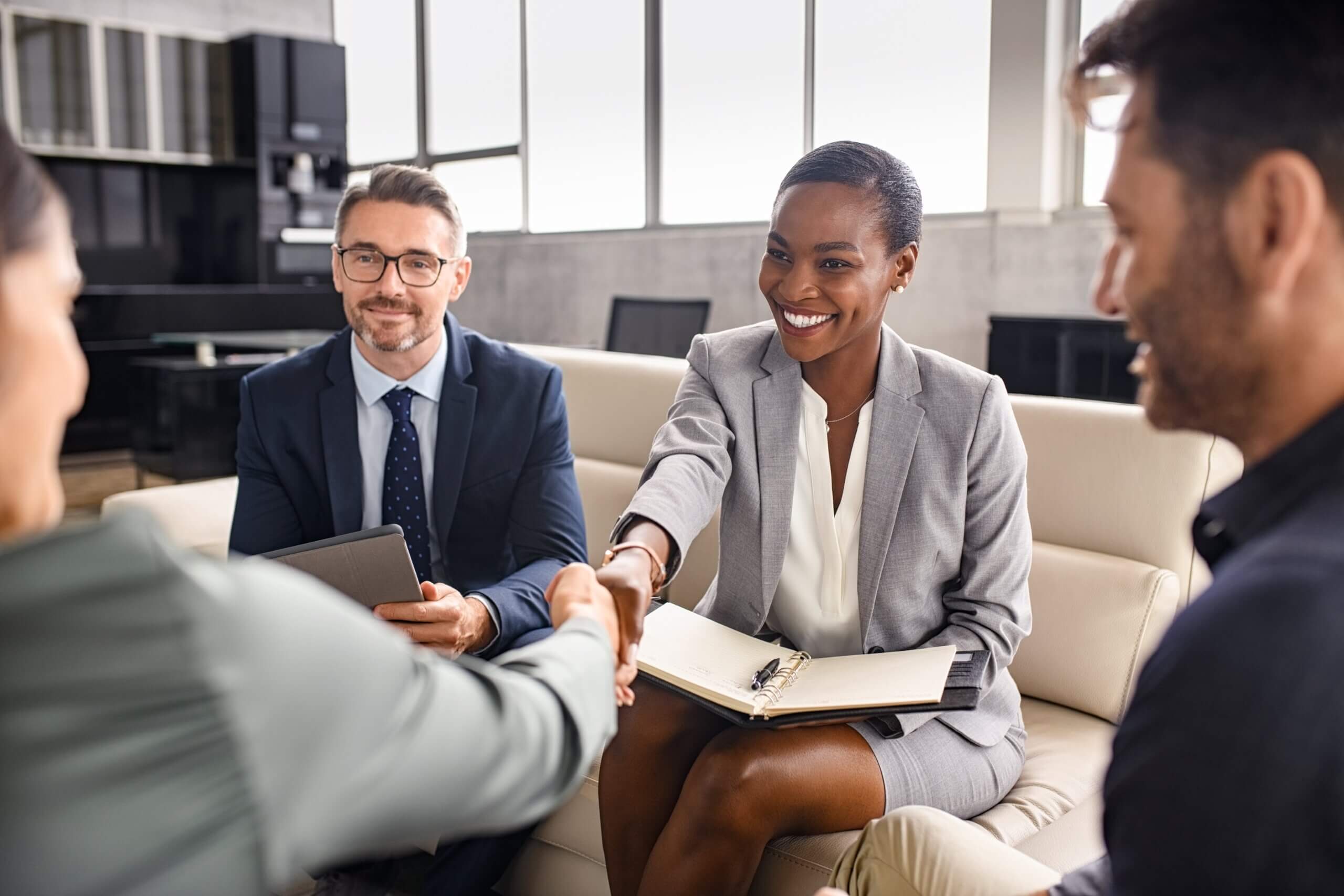 A group of four people in business attire are sitting on a couch. Two people in the center are shaking hands, expressing a professional agreement or meeting. One person holds a notepad and the others look engaged. The setting appears professional.