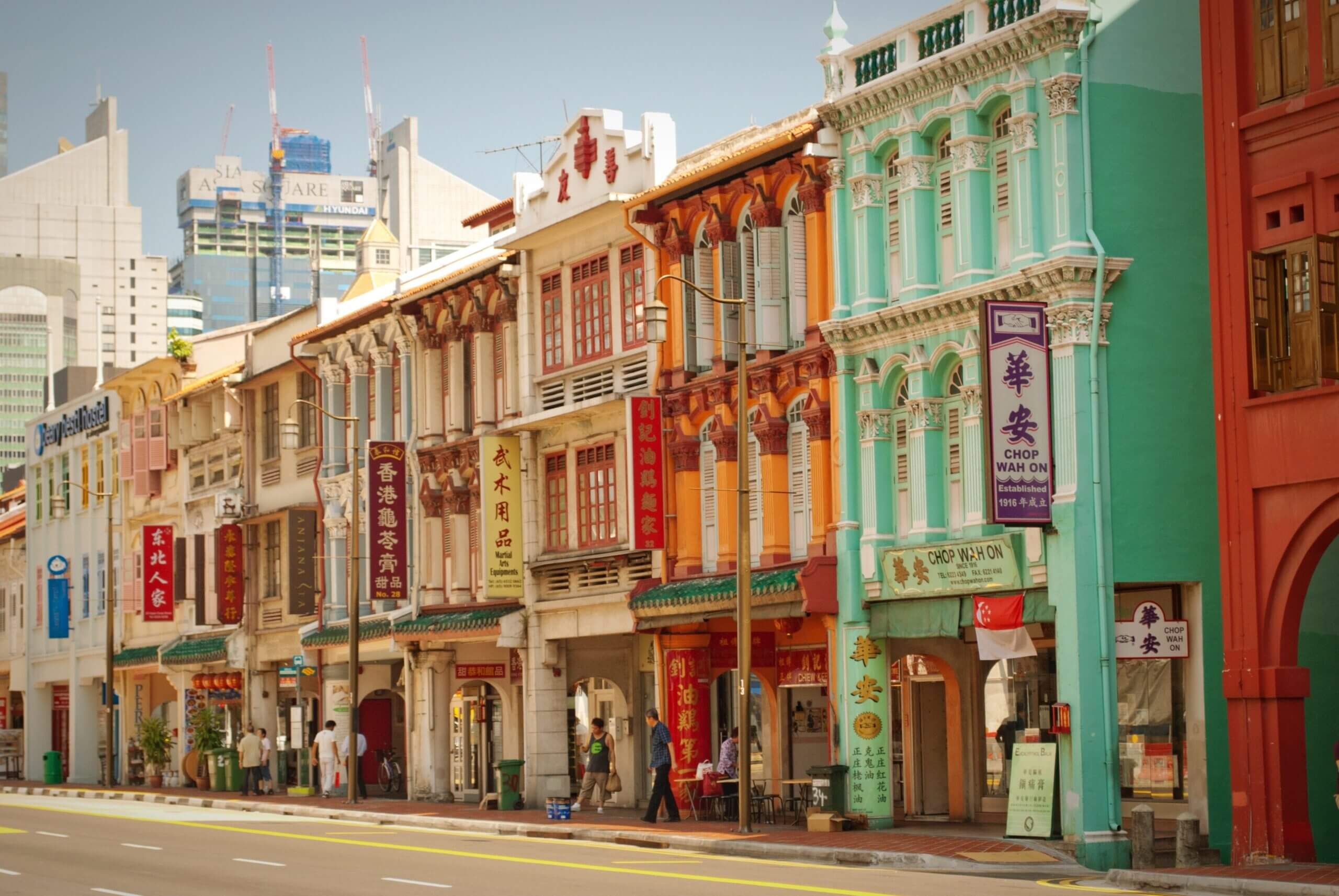A vibrant street lined with colorful, historic shophouses featuring ornate facades and decorative window shutters. Signs with Chinese characters hang from the buildings. Modern skyscrapers are visible in the background under a clear blue sky.