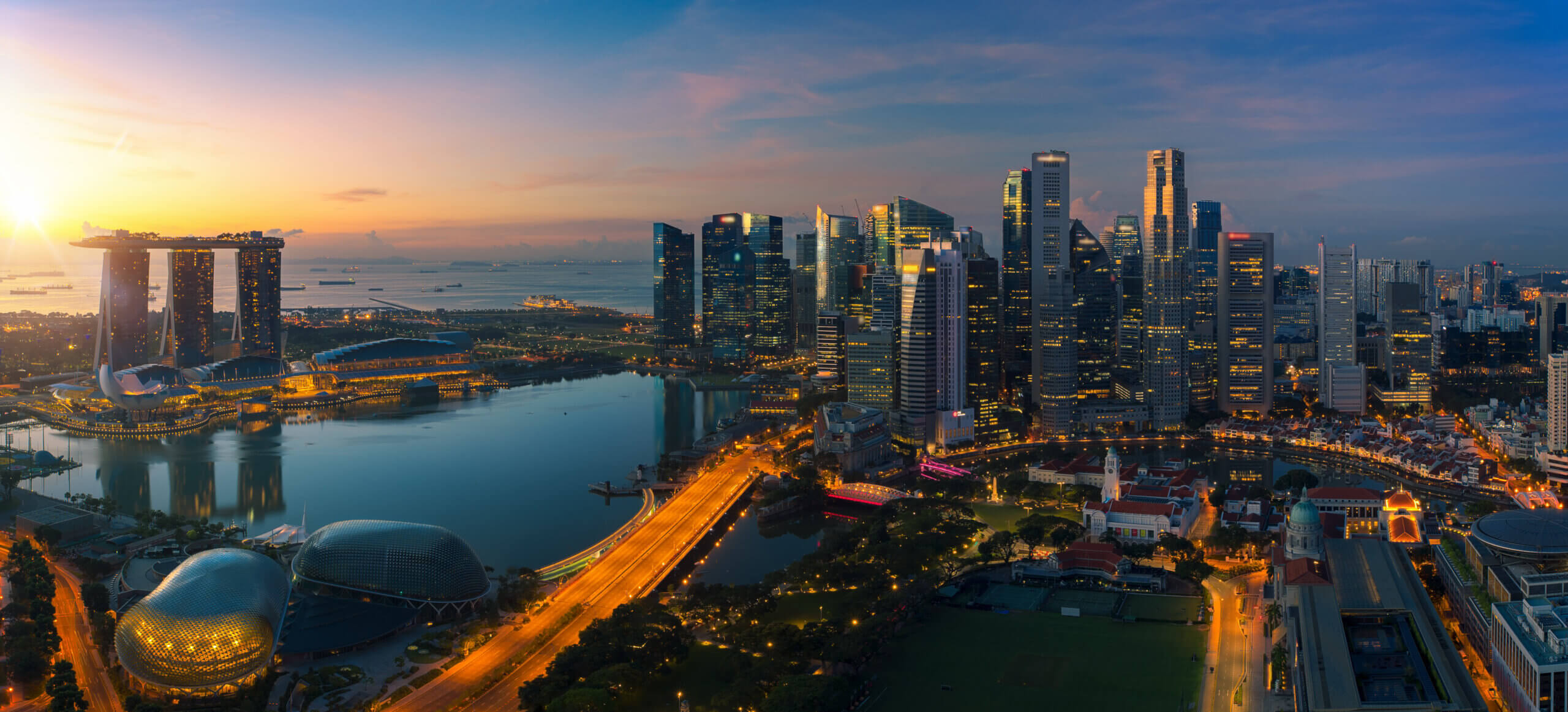 Aerial view of Singapore at sunrise, showcasing the Marina Bay Sands hotel and modern skyline with tall buildings. The sunlight reflects off the bay and structures, creating a vibrant urban scene with a clear sky in the background.