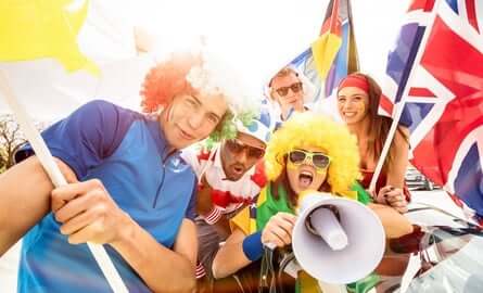 A group of five enthusiastic sports fans wearing colorful wigs and clothing, holding various international flags. One person is speaking into a megaphone. They appear excited and are smiling in a festive atmosphere.