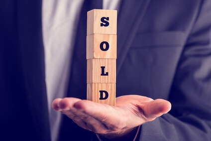 A person in a suit holding a stack of wooden blocks spelling "SOLD." The hand is open, palm facing up, supporting the vertically arranged blocks. The background is blurred, focusing attention on the blocks and hand.