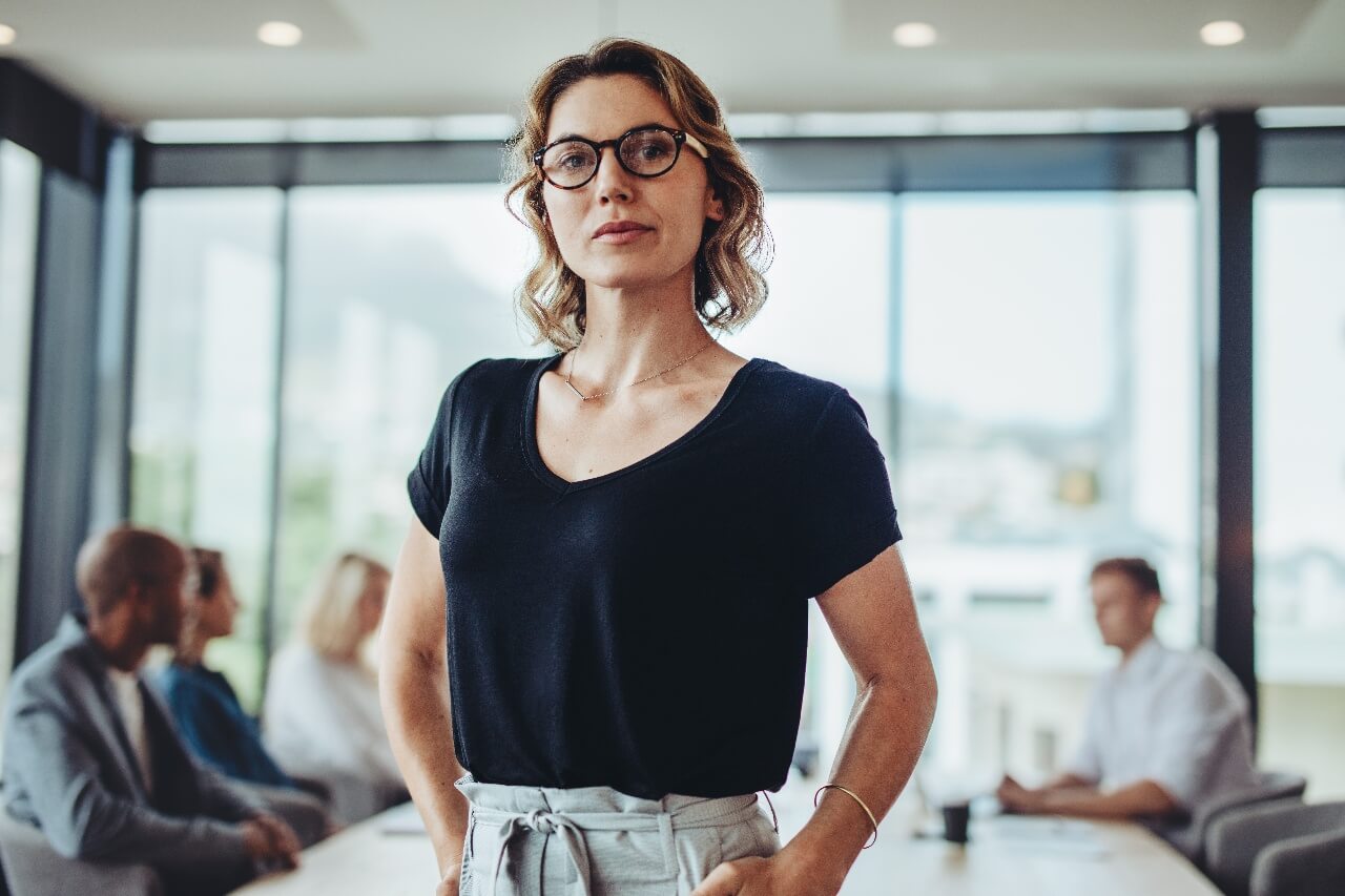 A woman with glasses stands confidently in an office room, wearing a black shirt and gray pants. In the background are four colleagues sitting at a table, engaged in conversation. Large windows provide natural light.