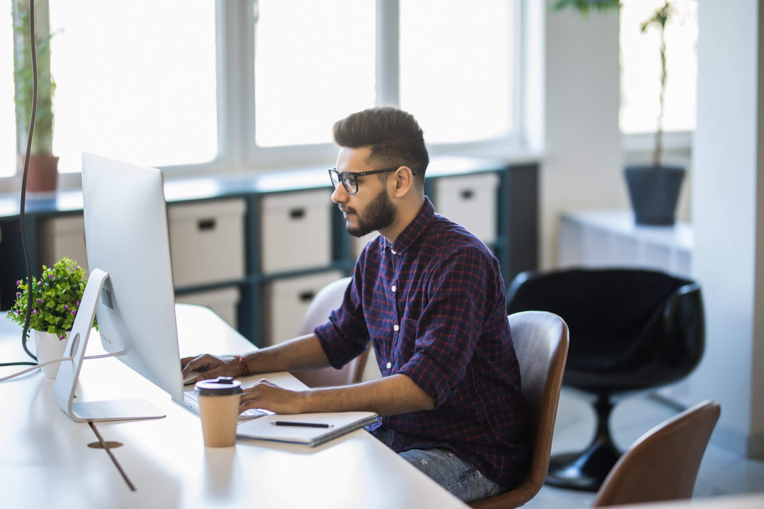 A bearded man in glasses works on a computer at a modern desk. He's wearing a plaid shirt and is focused on the screen. A notebook and a coffee cup sit nearby, with shelves and large windows in the background.