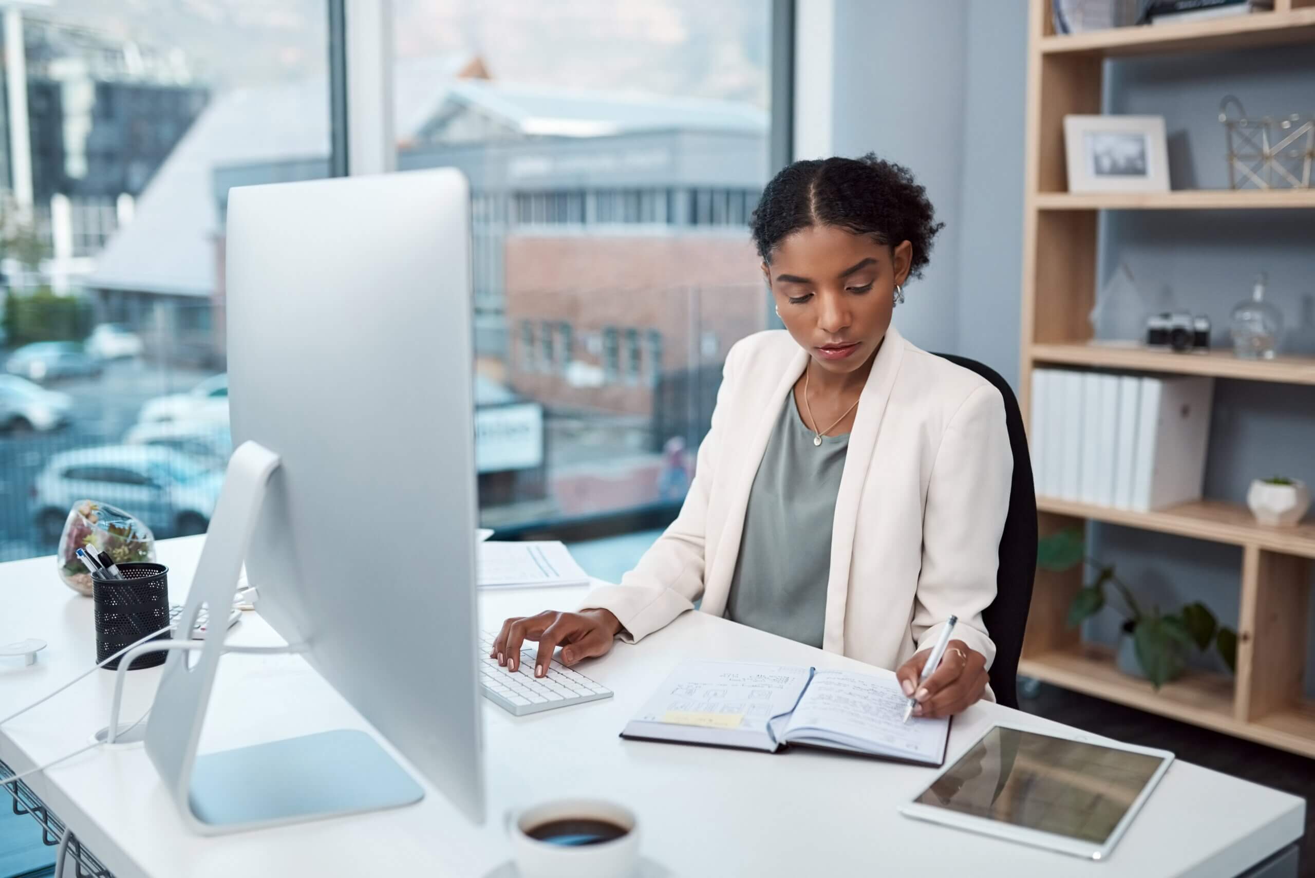 A woman in a white blazer sits at a desk in an office, writing in a notebook. She is working on a computer with a cup of coffee beside her. Large windows show a blurred view of buildings outside.