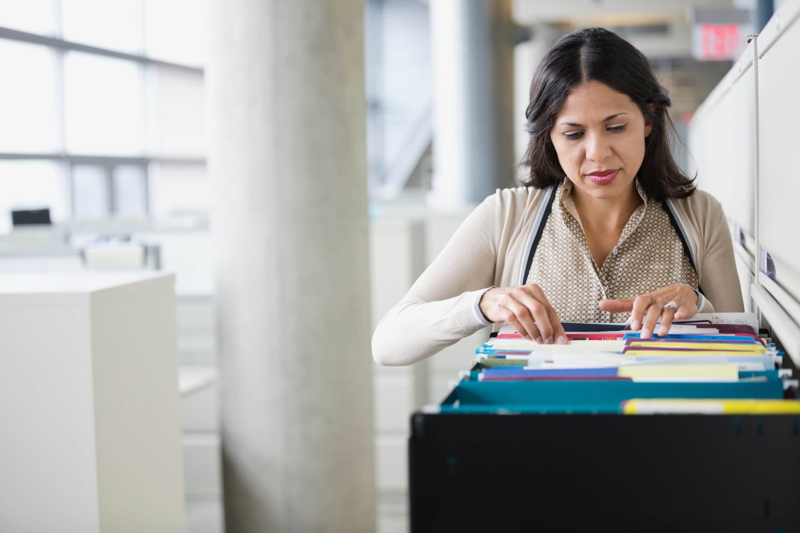 A woman in a cardigan and scarf is looking through files in an open filing cabinet drawer in a modern office space with large windows. She appears focused on her task.