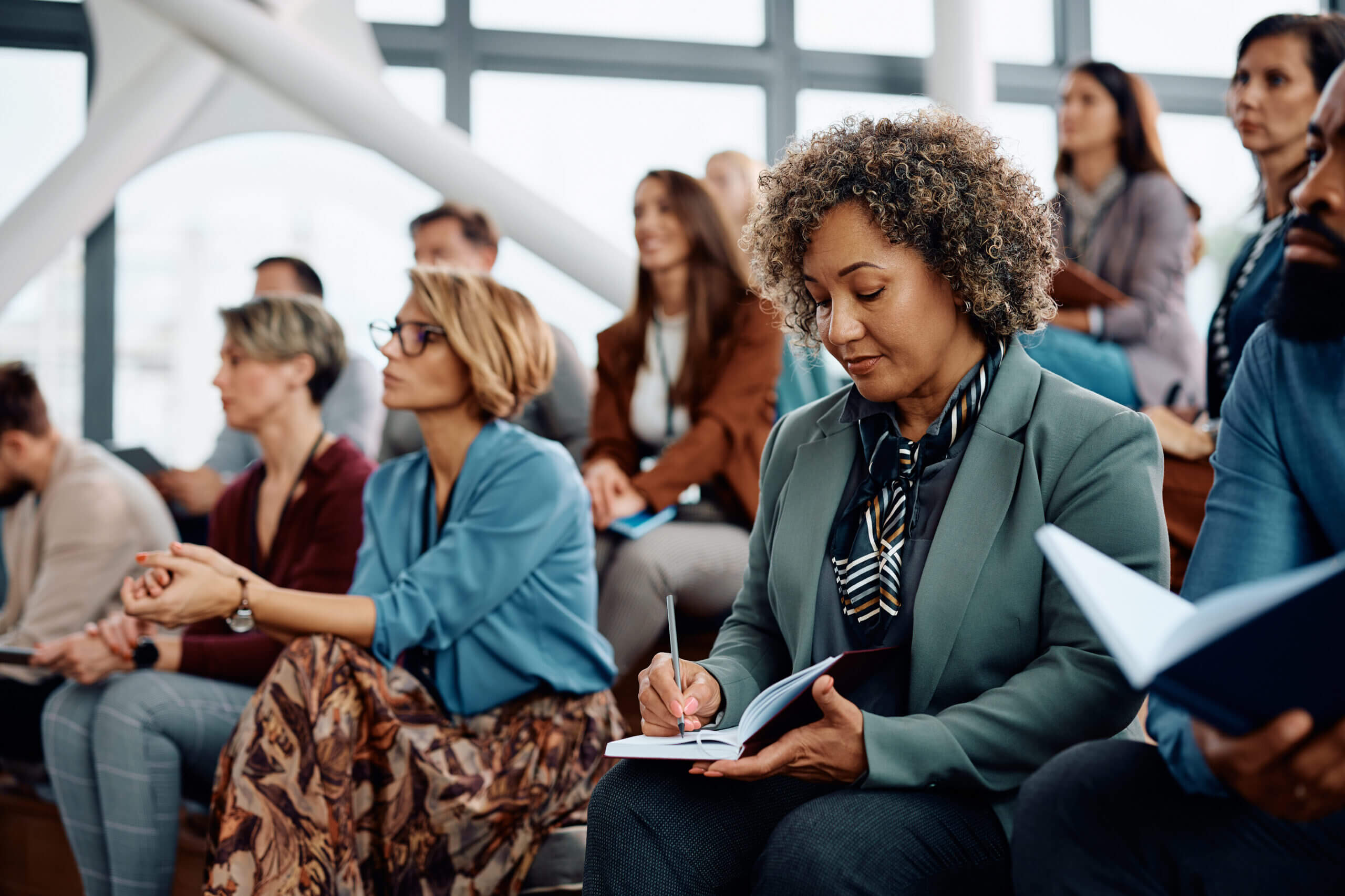 A diverse group of people is seated in a modern conference room. A woman with curly hair, wearing a green blazer, is writing notes in a notebook. Other attendees are engaged, some taking notes, with large windows in the background.