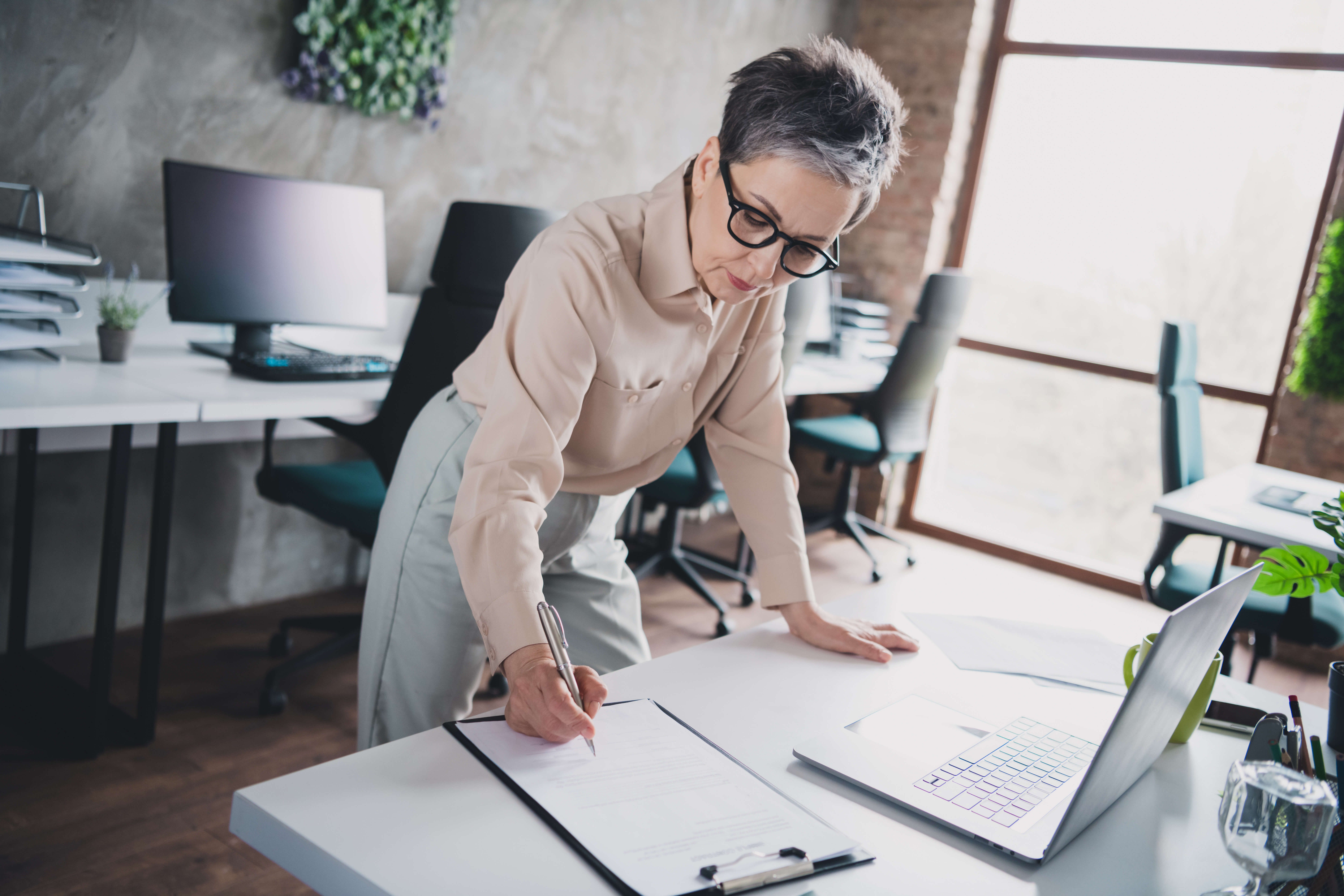 An older adult with short gray hair is standing at a desk in an office, signing a document on a clipboard. They are wearing glasses and a light-colored outfit. A laptop, potted plant, and office supplies are visible on the desk.