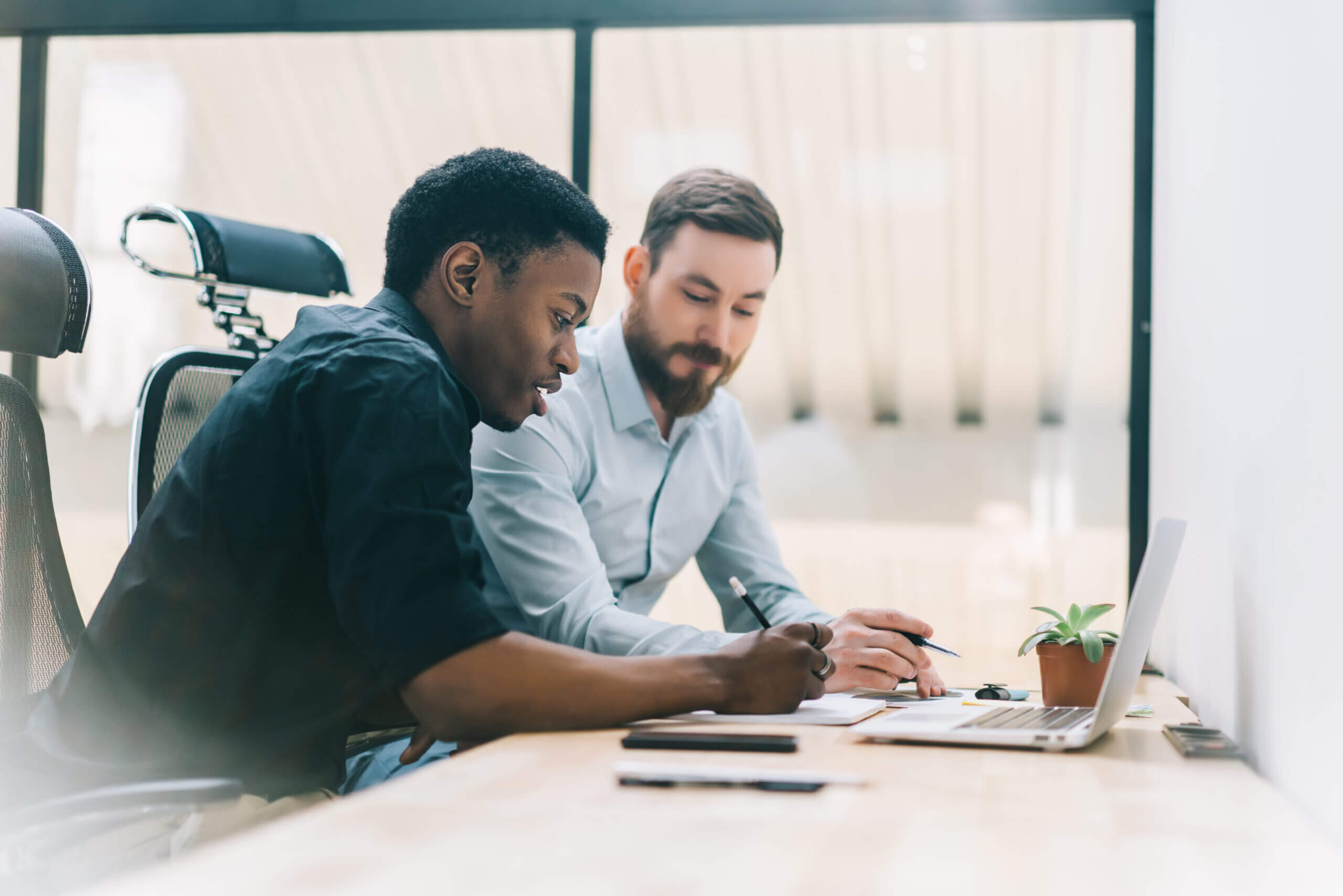 Two men collaborating in an office environment. One is writing notes on paper, while the other observes. A laptop, phone, and small plant are on the desk. Large windows provide natural light.