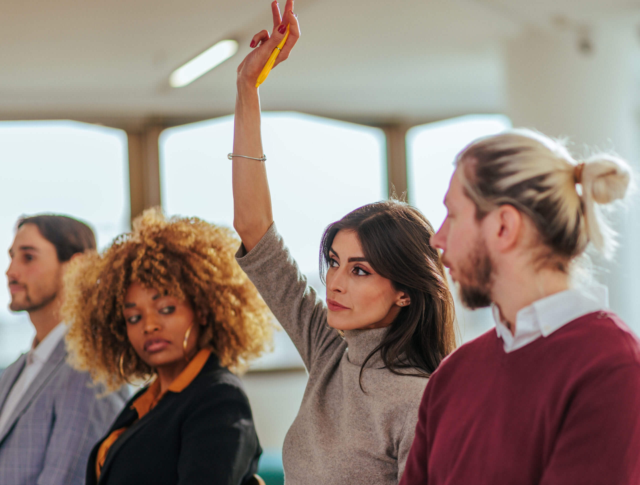 A woman in a gray sweater with long hair raises her hand in a group setting. She is seated between a woman with curly hair and a man with a bun. Another man in a suit is visible. They appear to be in a meeting or class.