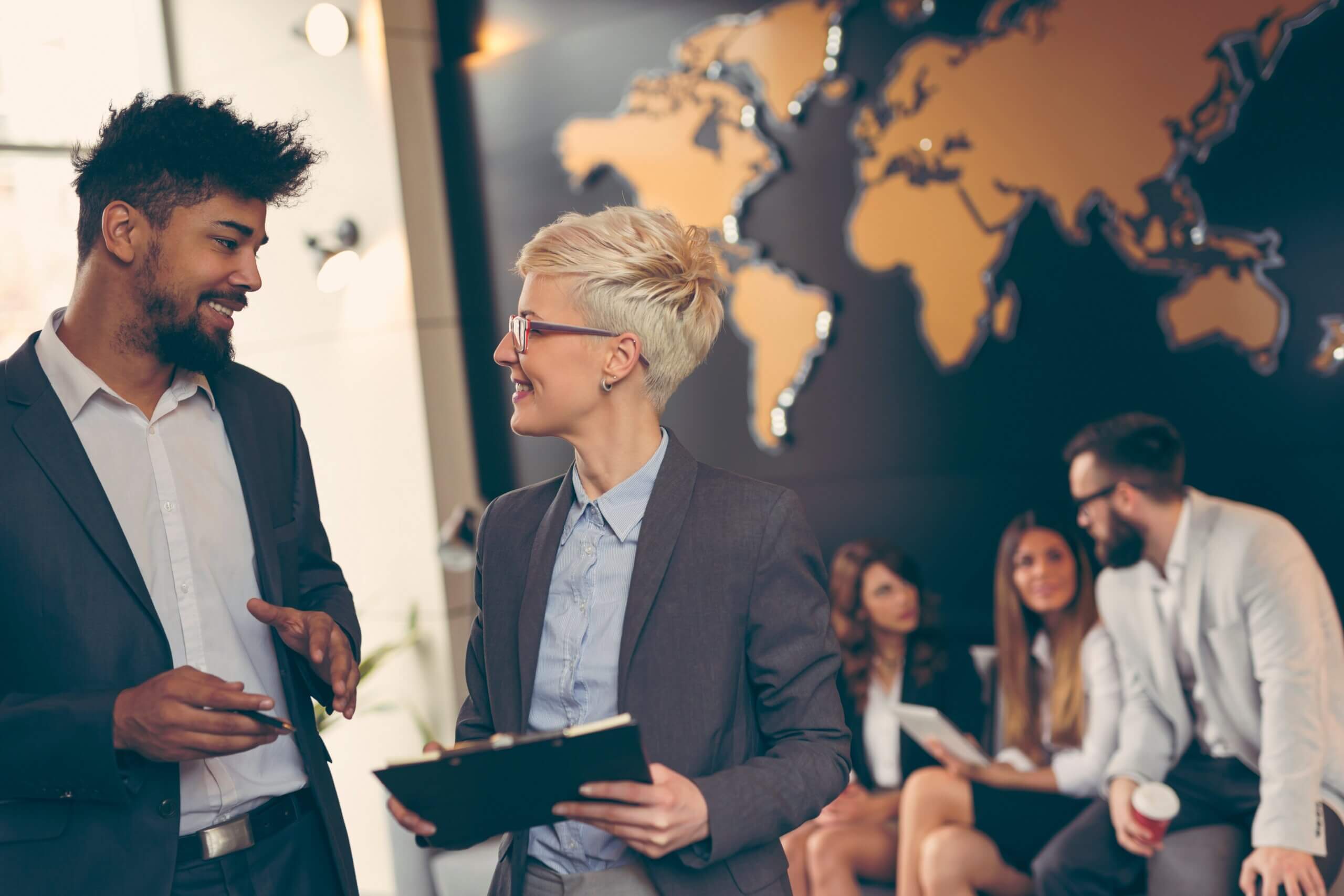 A man and woman in business attire converse in an office with a world map on the wall. The woman holds a clipboard. In the background, three people in business clothes sit together, engaging in conversation.