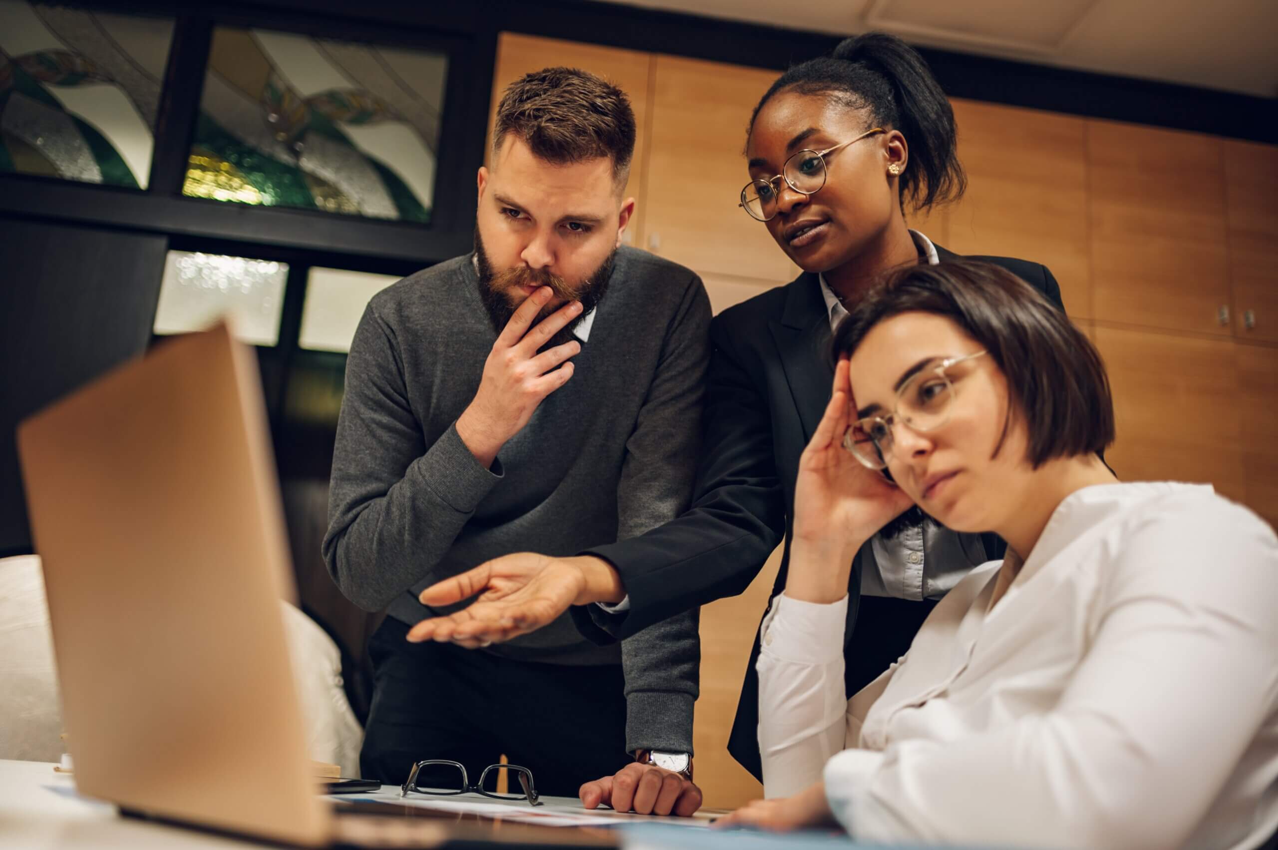 Three people gathered around a laptop in an office setting. A man with a beard is explaining something, while a woman in a blazer listens attentively. Another woman in glasses is seated, looking thoughtfully at the laptop screen.